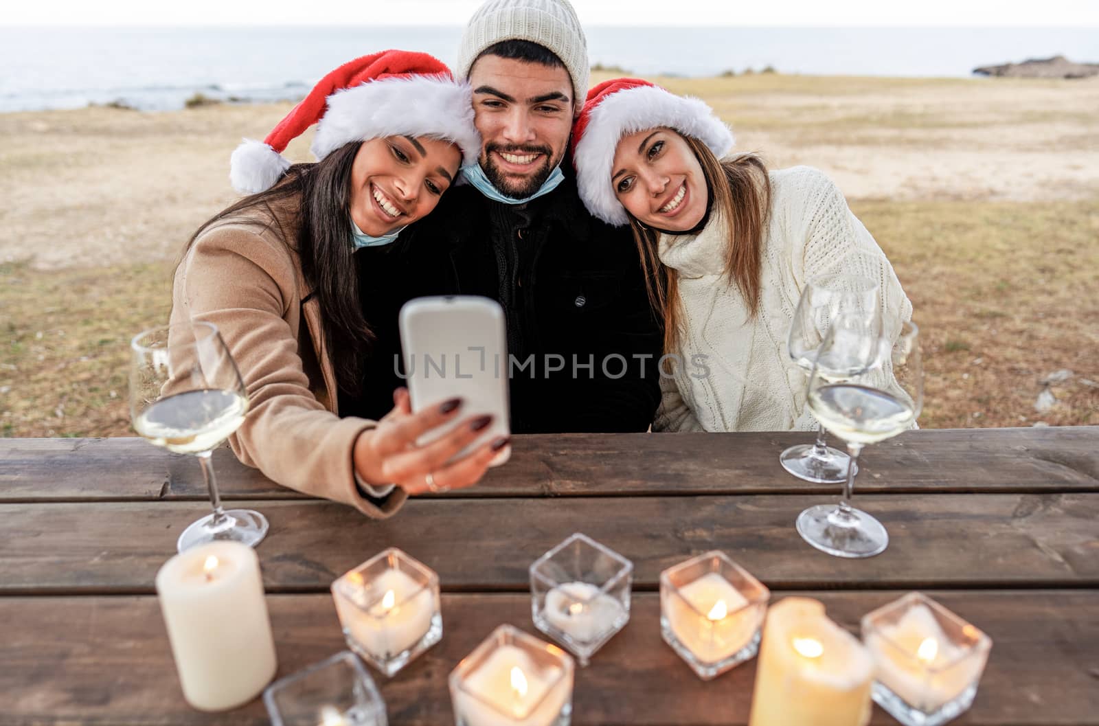 Three mixed race friends with lowered mask sitting on a wooden table outdoor in sea resort celebrating Christmas and New Year event making selfie with wine glasses - Smiling women posing in Santa hat