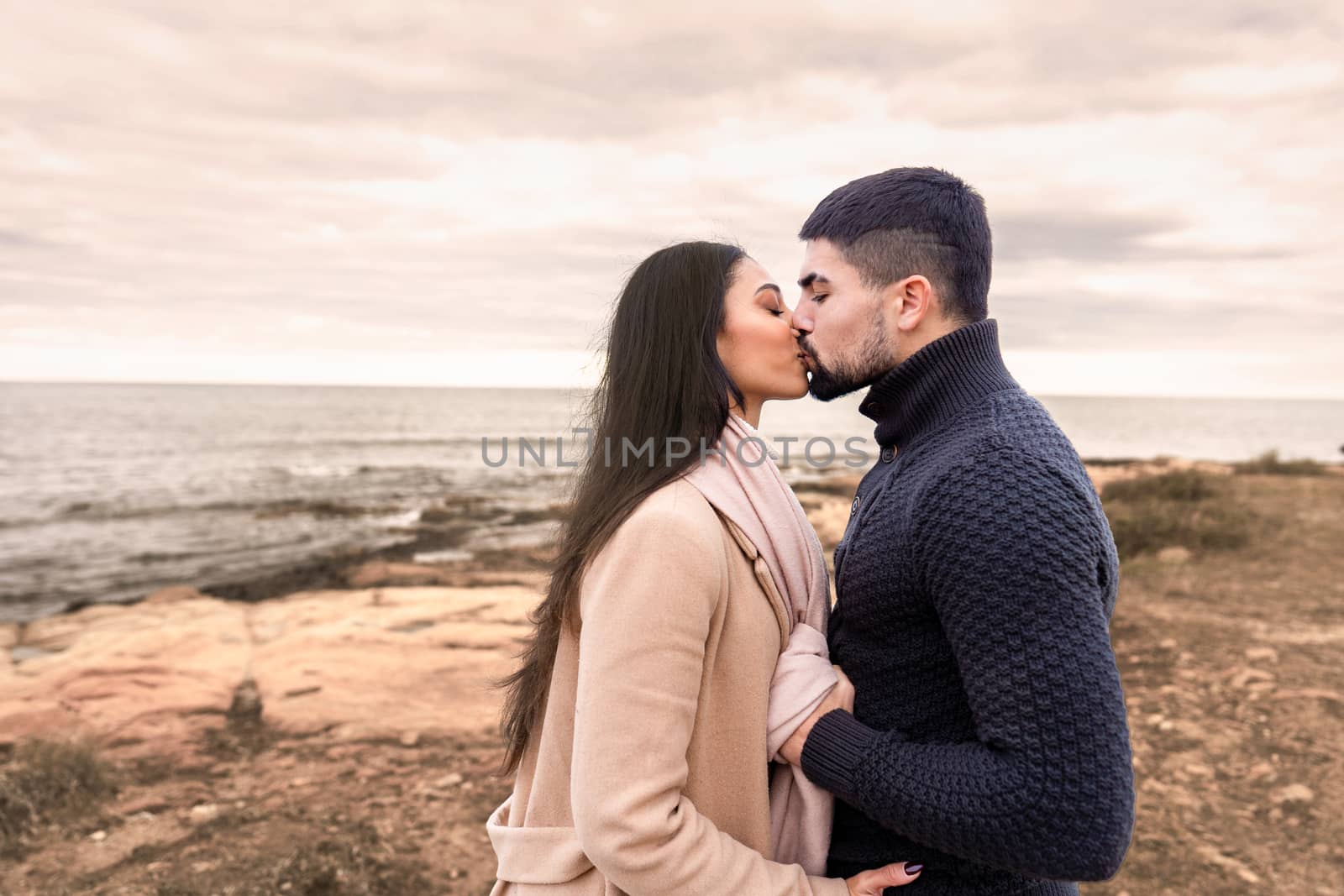 Mixed race couple romance scene on sea rocks and cloudy sky at sunset or dawn - Handsome bearded guy kisses his beautiful black Hispanic long hair woman holding her by the scarf with autumn colors by robbyfontanesi