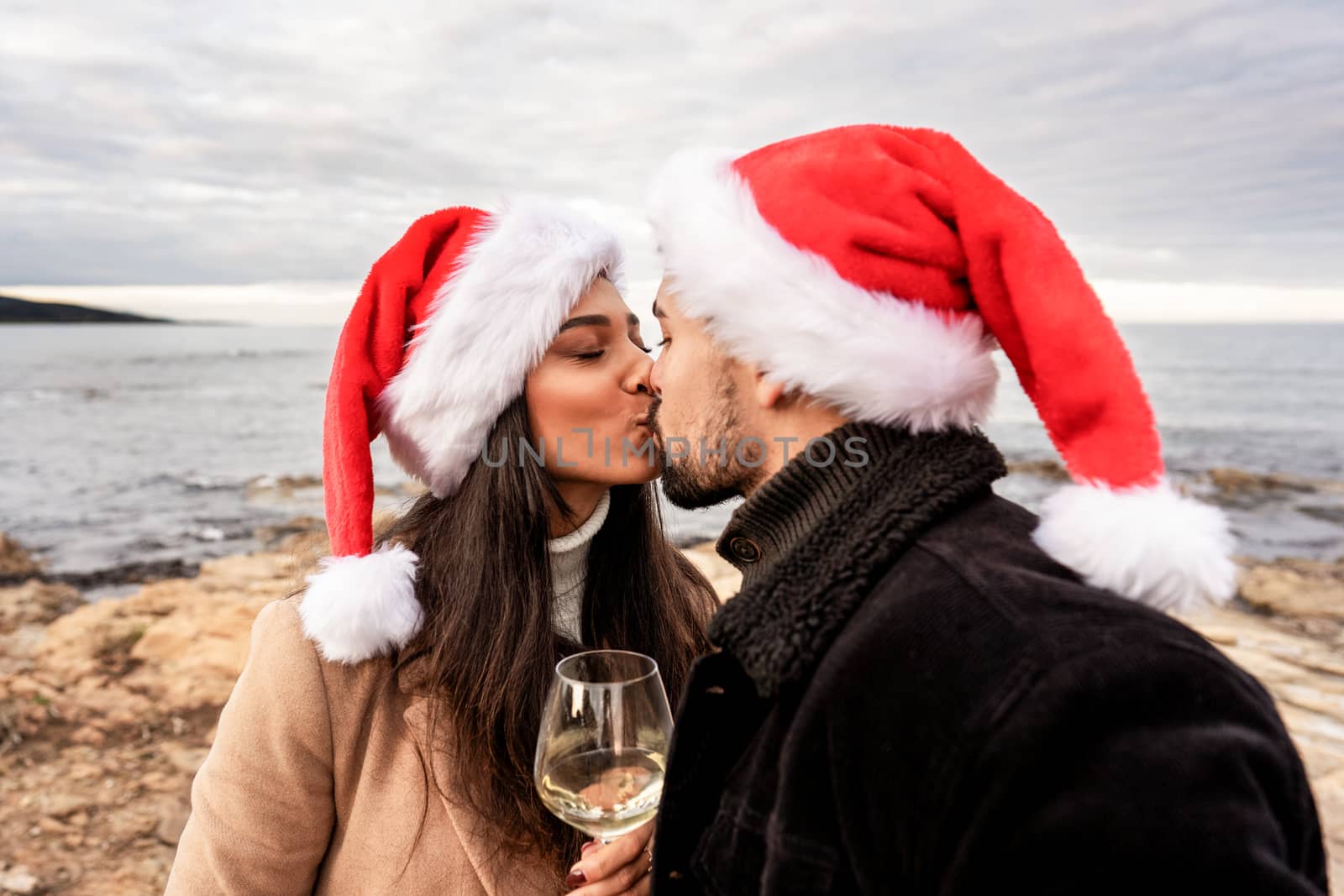 Passionate kiss of lover couple on sea rocks in winter sea Christmas and New Year vacation - Young beautiful girlfriend and boyfriend bonding wearing Santa hat holding wine glass at sunset on ocean