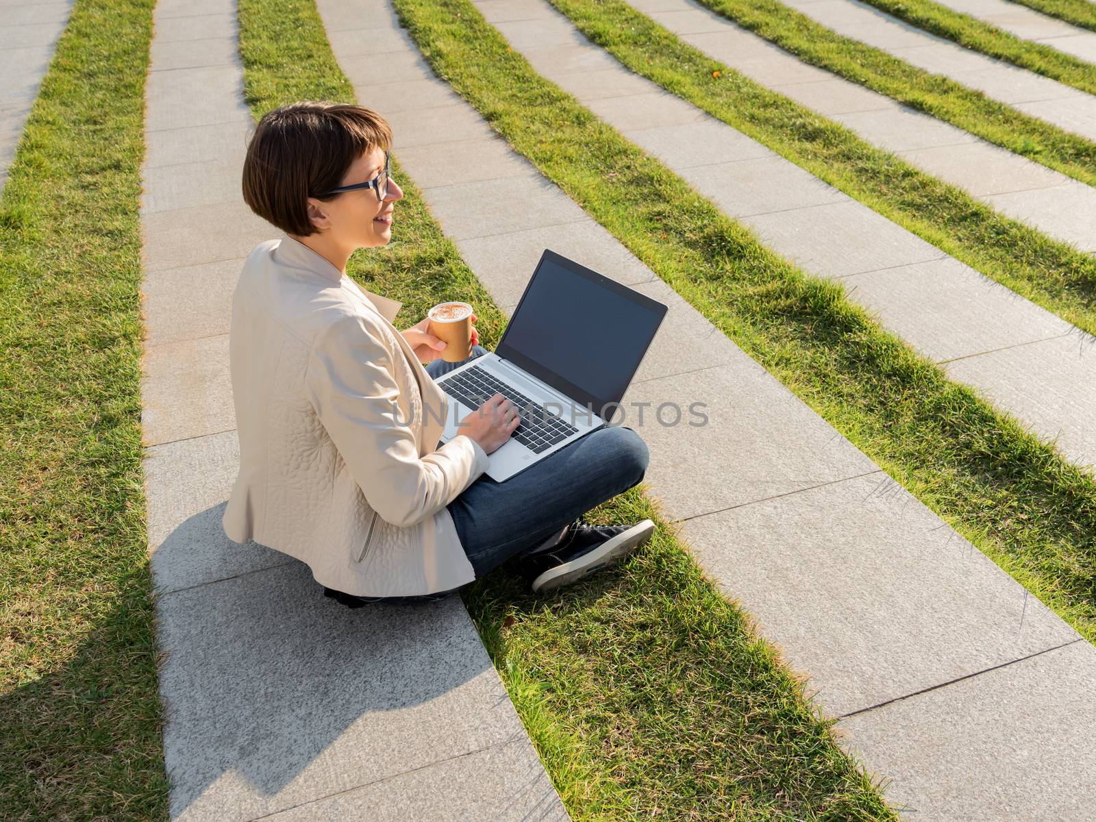 Freelance business woman sits in park with laptop and take away cardboard cup of coffee. Casual clothes, urban lifestyle of millennials. Working remotely.