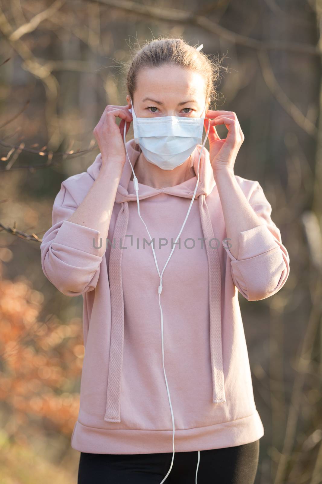 Portrait of caucasian sporty woman wearing medical protection face mask while walking in park, relaxing and listening to music. Corona virus, or Covid-19, is spreading all over the world.