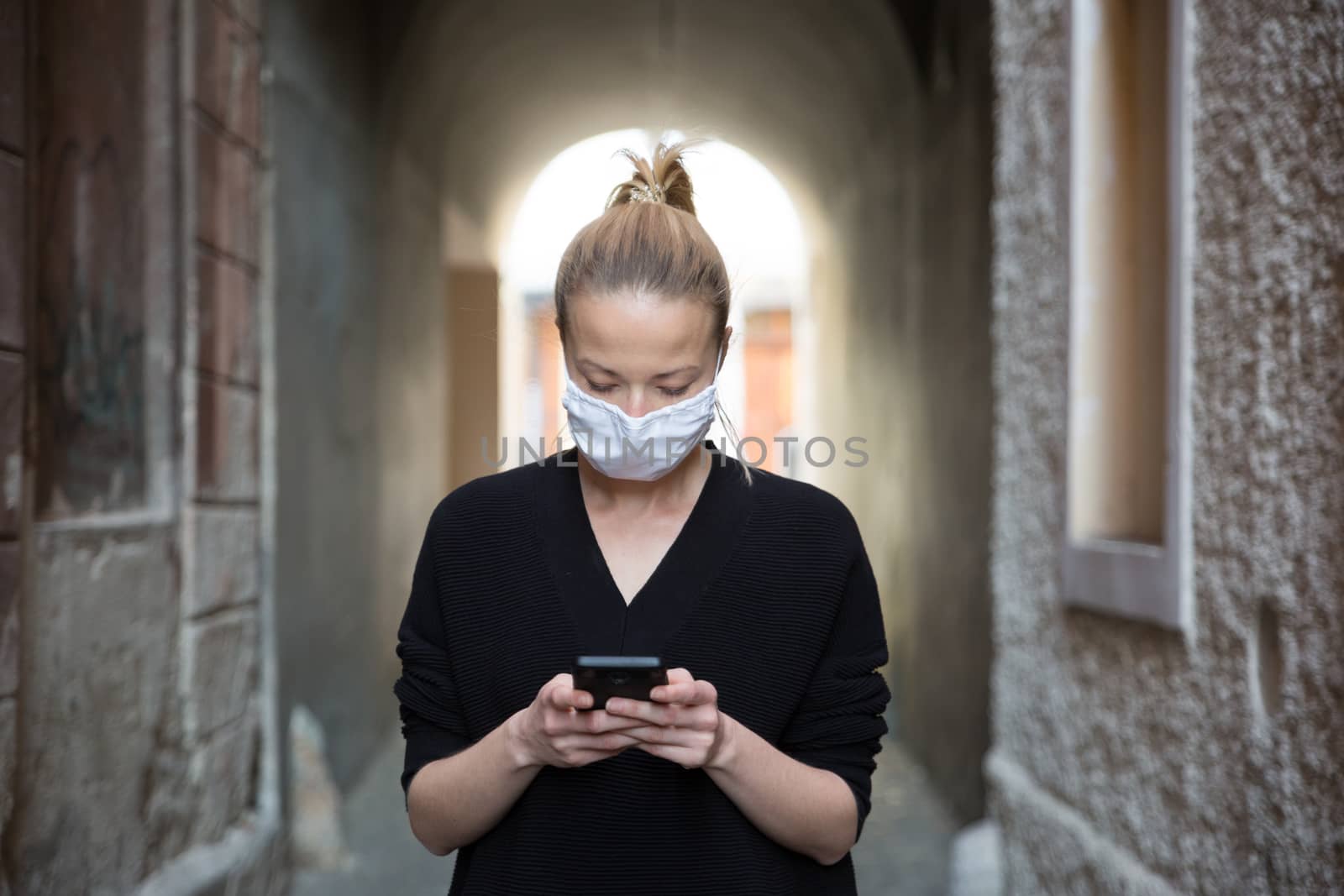 COVID-19 pandemic coronavirus. Casual caucasian woman at medieval city street using mobile phone, wearing protective face mask against spreading of coronavirus and disease transmission.
