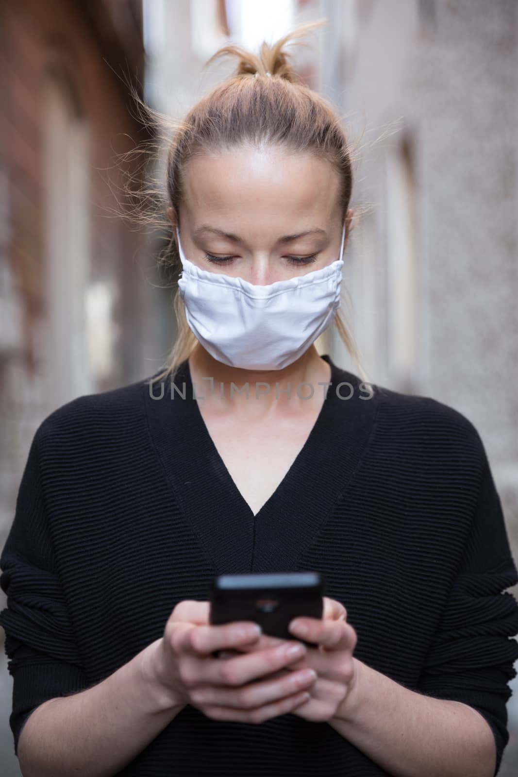 COVID-19 pandemic coronavirus. Casual caucasian woman at medieval city street using mobile phone, wearing protective face mask against spreading of coronavirus and disease transmission.