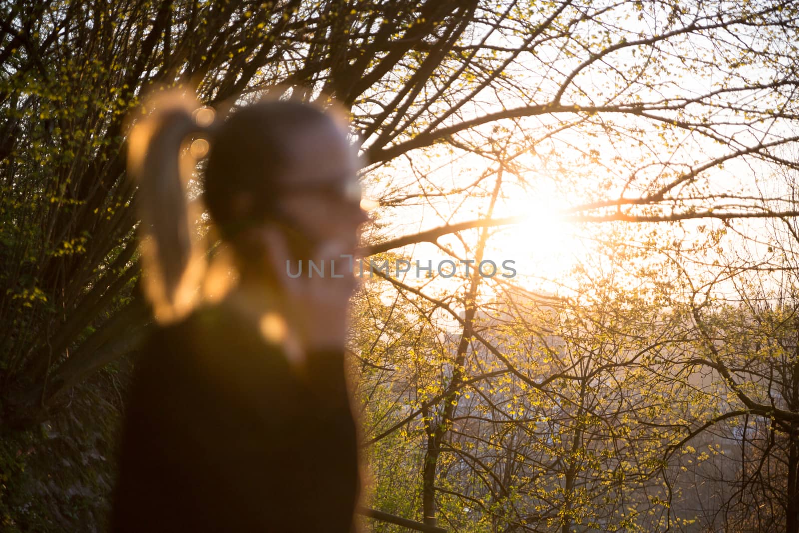 Backlit defocused rear view of young woman talking on cell phone outdoors in park at sunset. Girl holding mobile phone, using digital device, looking at setting sun.