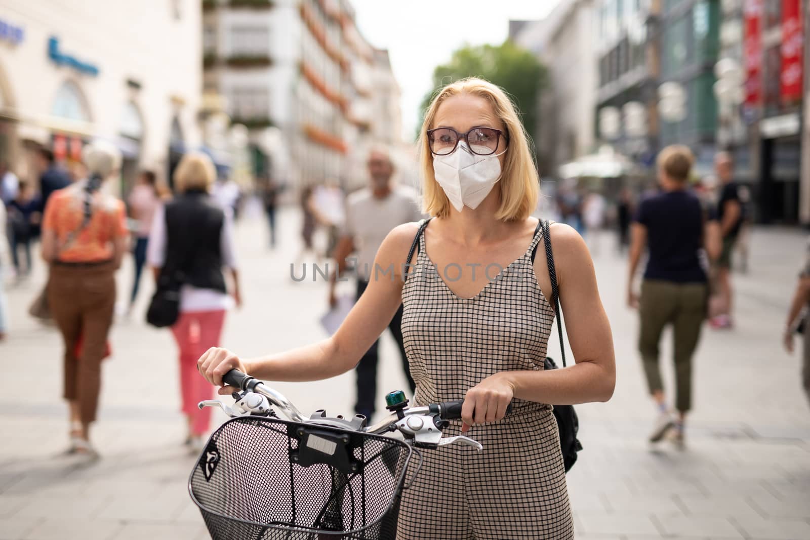 Woman walking by her bicycle on pedestrian city street wearing medical face mask in public to prevent spreading of corona virus. New normal during covid epidemic. Social responsibility.