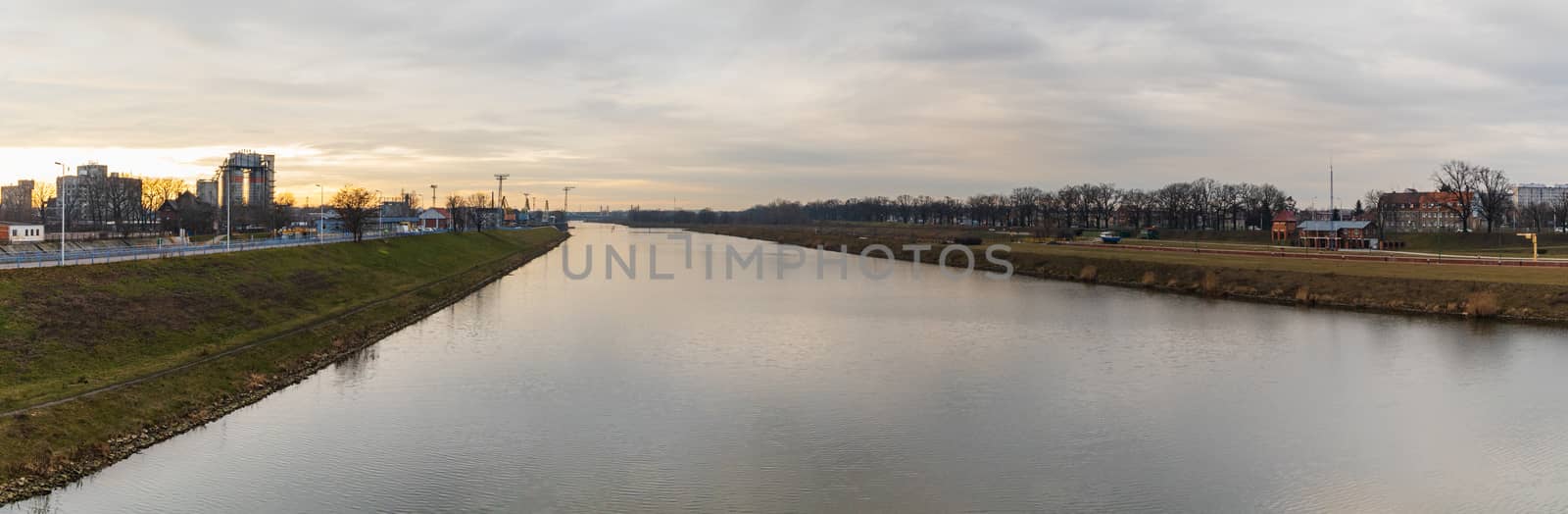 Cloudy panorama of Odra river between green coasts of city at sunset by Wierzchu