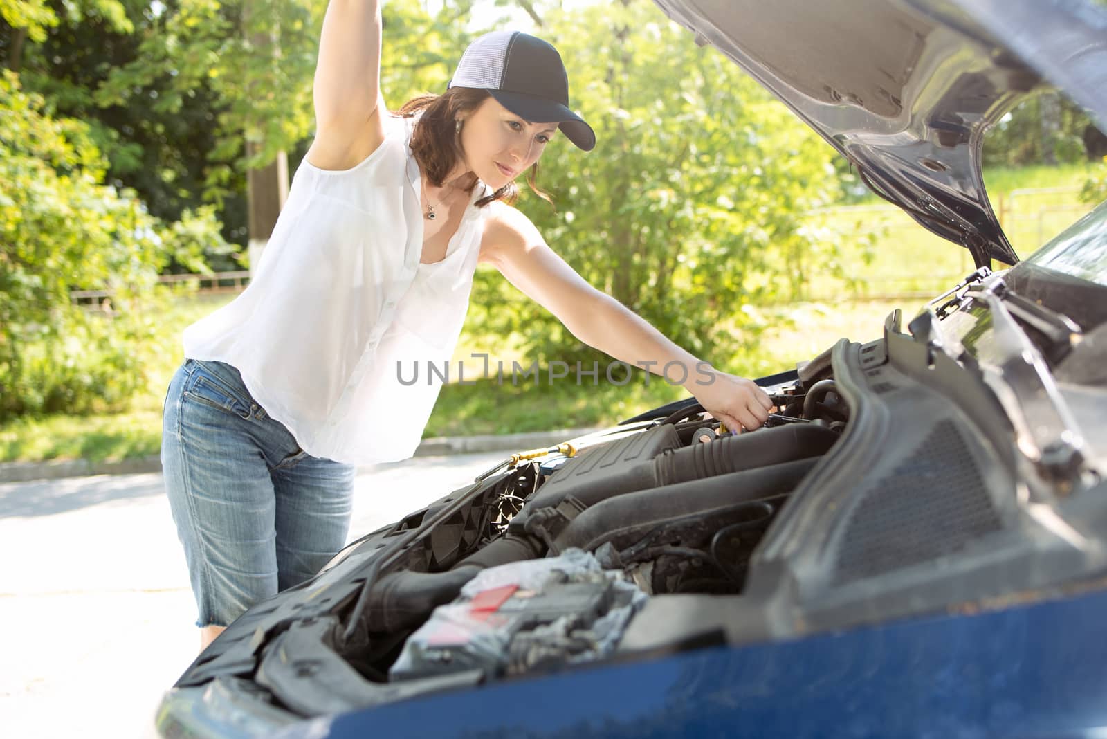 A woman in hat and denim shorts waits for assistance near her car broken down on the road side. by Nickstock