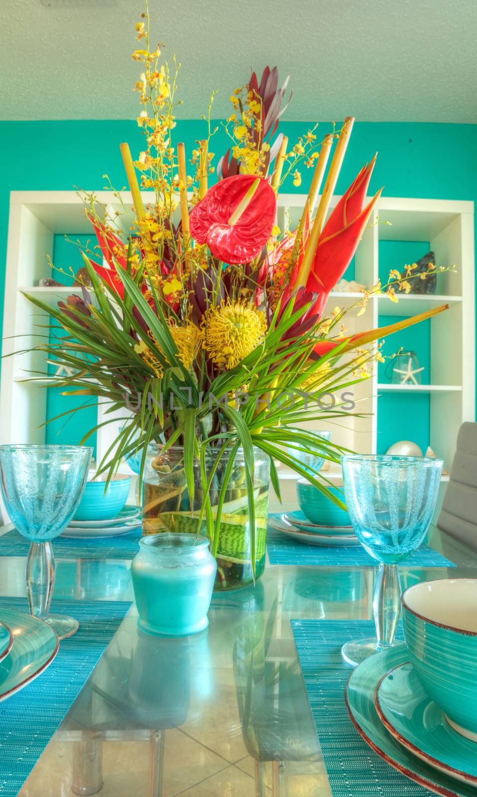 Tropical blue table set with flowers including Heliconia bihai, yellow oncidium orchids, yellow pincushion protea, and red anthuriums.