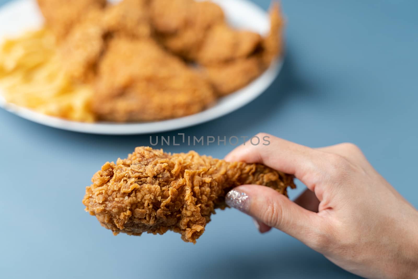 Hand holding drumsticks, crispy fried chicken with french fries in white plate on blue background.