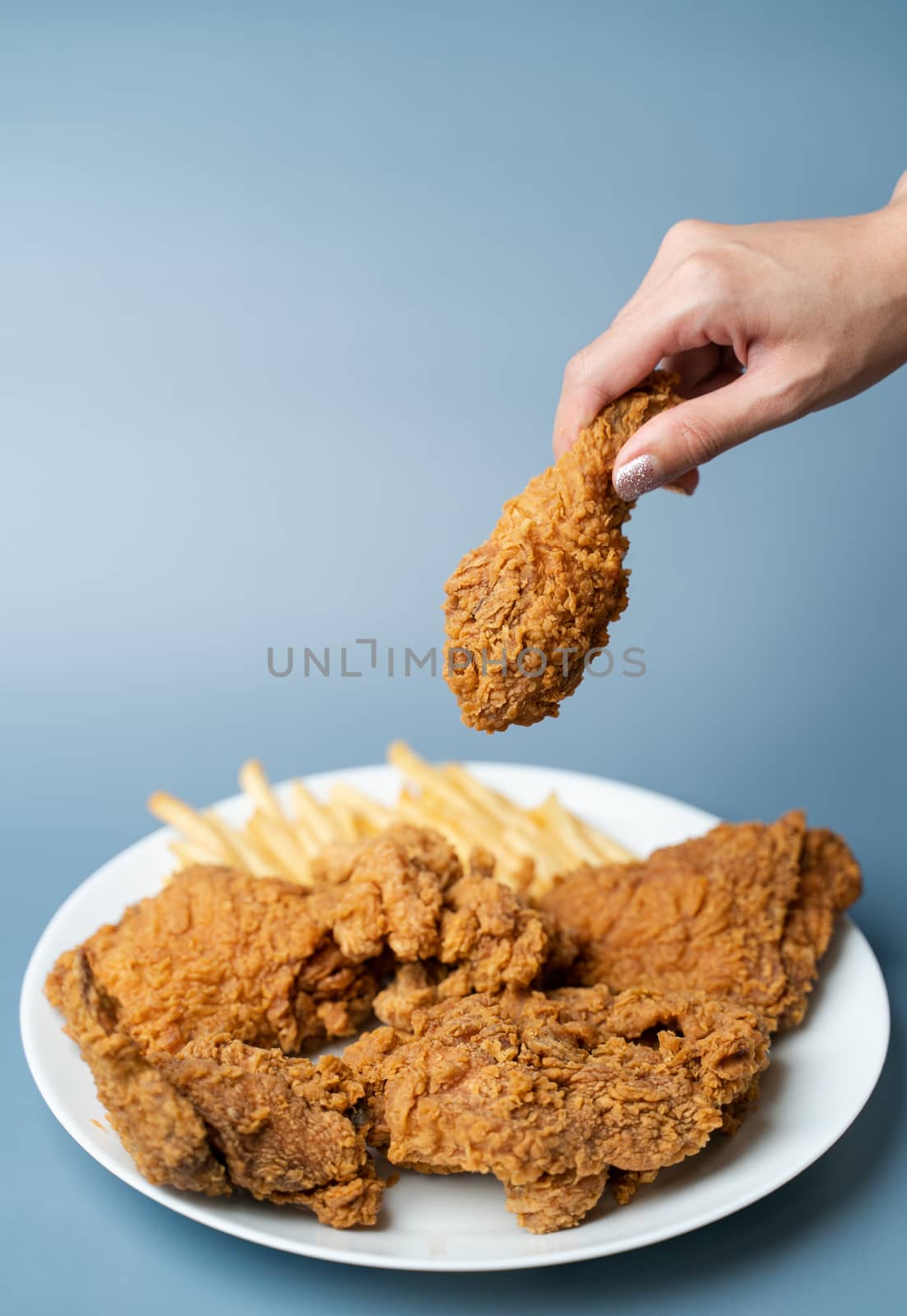 Hand holding drumsticks, crispy fried chicken with french fries in white plate on blue background.