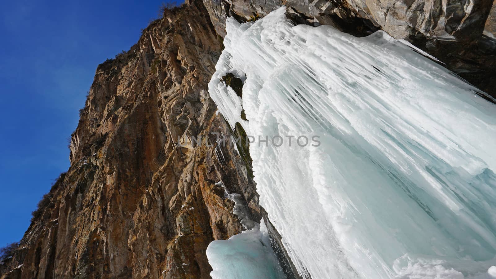 Frozen waterfall among the rocks. The waterfall is freezing, huge icicles. Ice white and blue. Brown rocks and splashes of water. Winter waterfall. White snow and blue sky. Water runs down the ice.