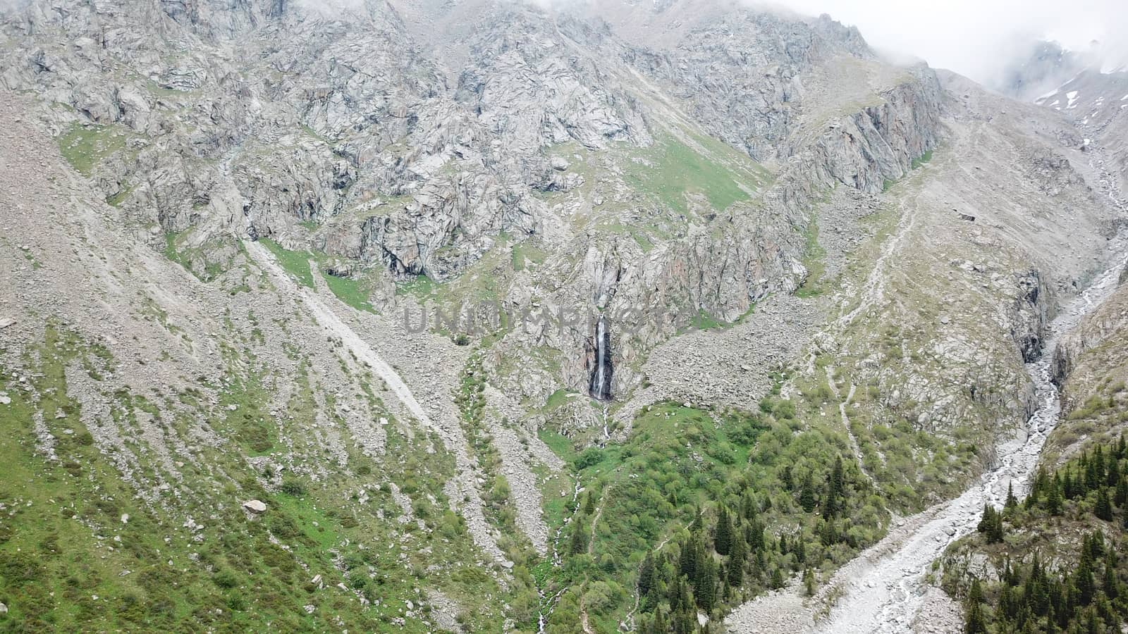 High waterfall among the rocks. View from a drone. Large rocks and steep slopes surround the mountain waterfall. Trees and grass grow below. Clouds float by. The view from top. Ala Archa, Kyrgyzstan.