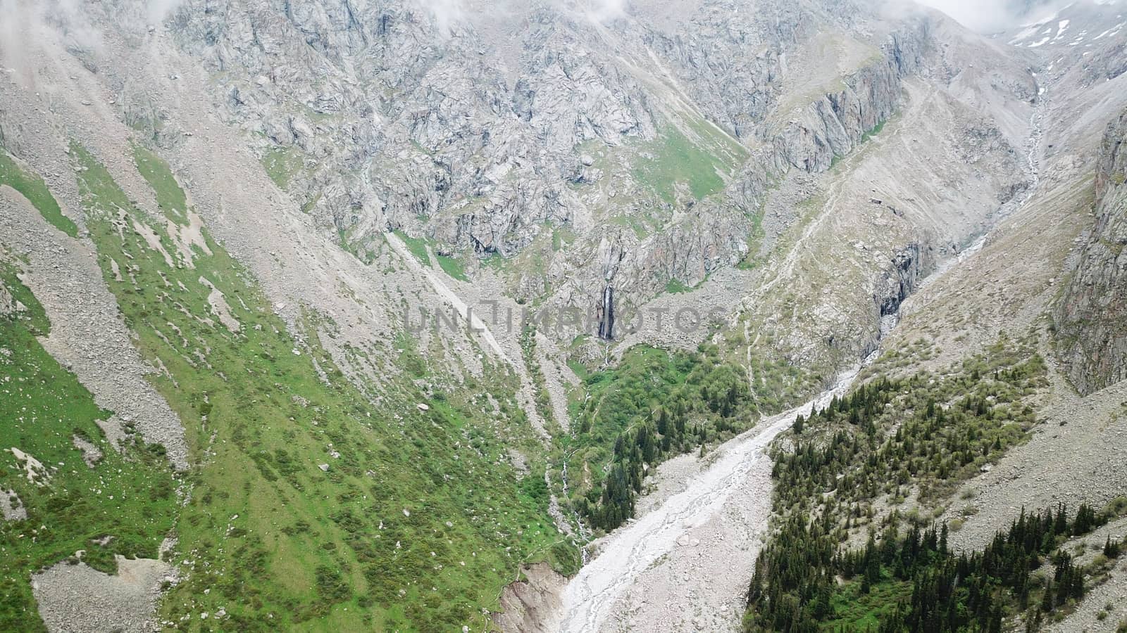 High waterfall among the rocks. View from a drone. Large rocks and steep slopes surround the mountain waterfall. Trees and grass grow below. Clouds float by. The view from top. Ala Archa, Kyrgyzstan.