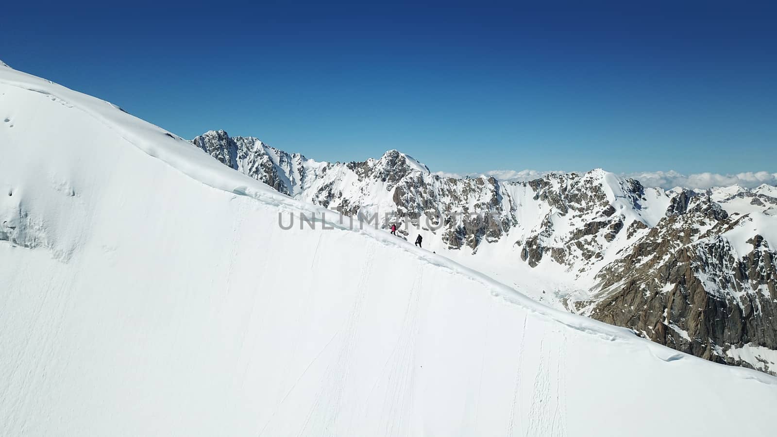 A group of climbers climb to the top of the peak. Dangerous, steep slope. Avalanche-prone area. Mountains covered with snow. Steep cliffs and large rocks. Above the mountains, top view from a drone.