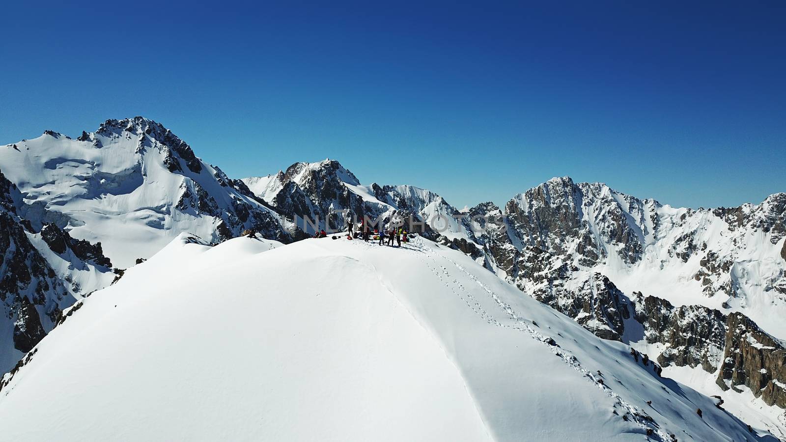 A group of climbers climb to the top of the peak. Dangerous, steep slope. Avalanche-prone area. Mountains covered with snow. Steep cliffs and large rocks. Above the mountains, top view from a drone.