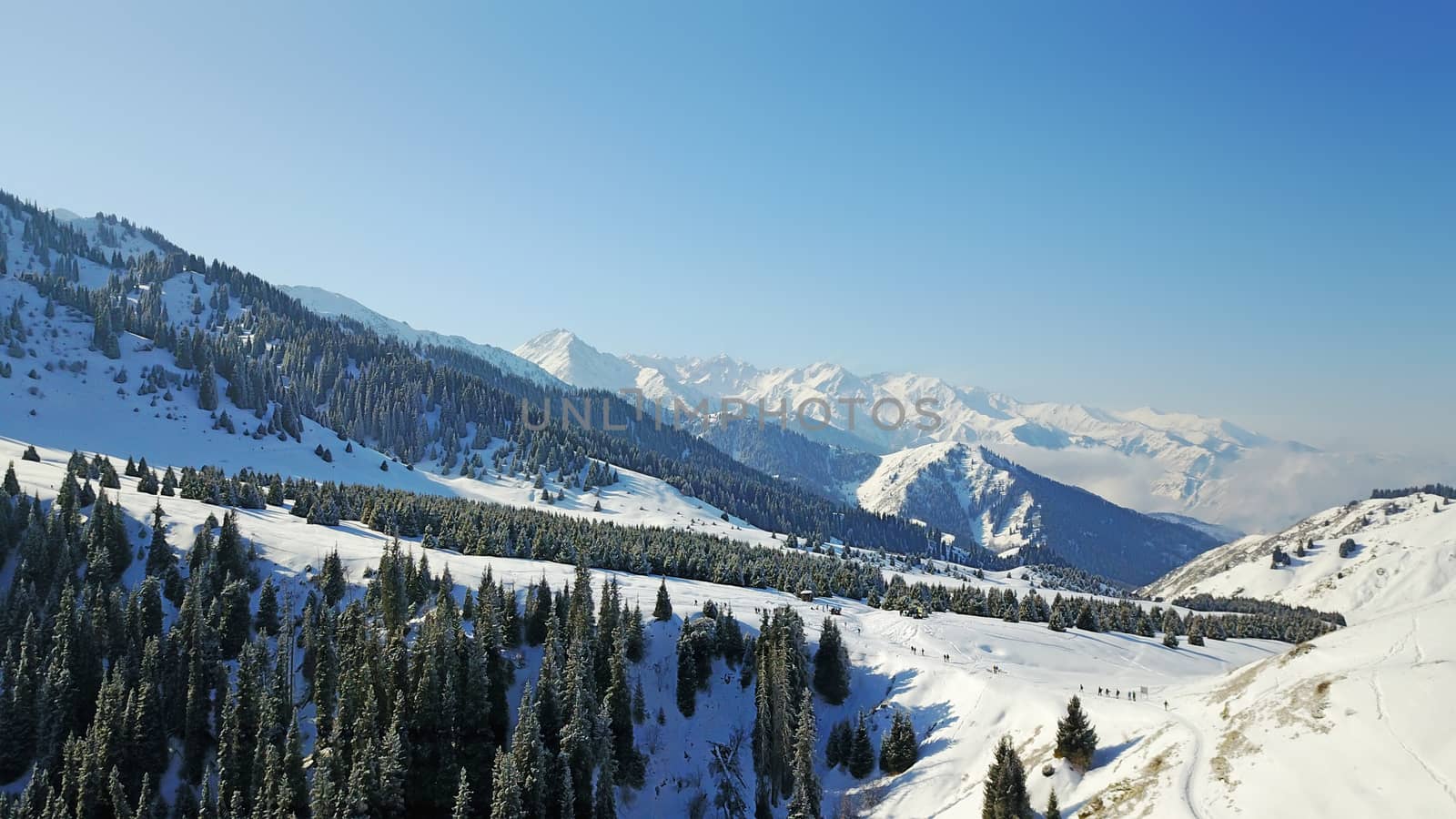 View from the height of the snowy mountains and forest. Huge clouds cover the hill in the distance. People are visible in places. Someone is running, resting, having a picnic or climbing to the top.