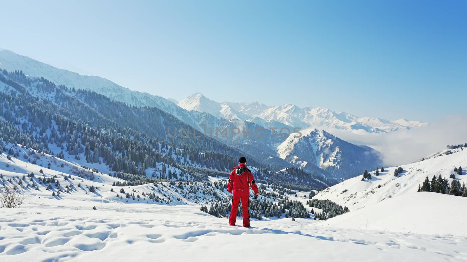 The guy looks at the snow hills and clouds. Mountainous terrain, all covered with fresh snow. Blue sky and huge clouds float along the gorge. On the slopes of a small forest, coniferous trees.