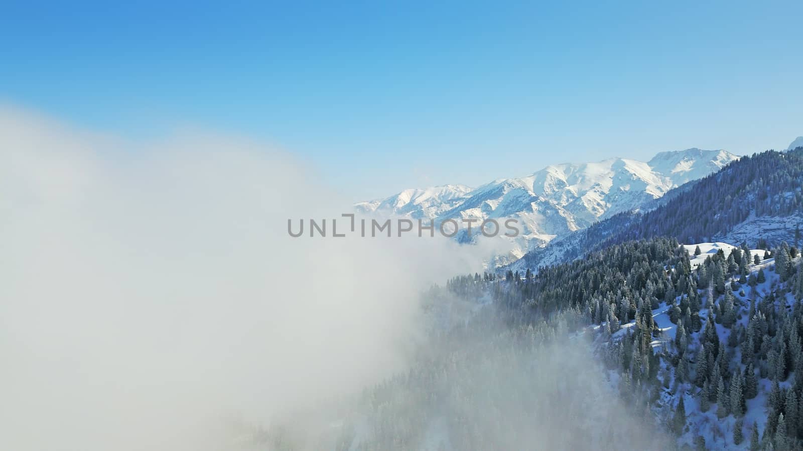 View from the height of the snowy mountains and forest. Huge clouds cover the hill in the distance. People are visible in places. Someone is running, resting, having a picnic or climbing to the top.