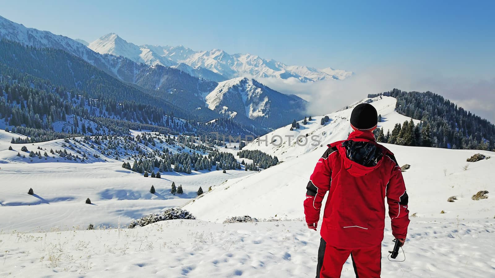 The guy looks at the snow hills and clouds. Mountainous terrain, all covered with fresh snow. Blue sky and huge clouds float along the gorge. On the slopes of a small forest, coniferous trees.