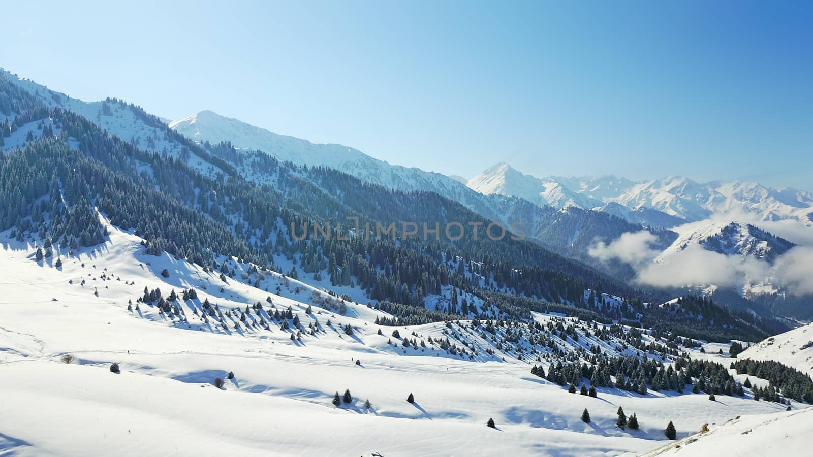 View from the height of the snowy mountains and forest. Huge clouds cover the hill in the distance. People are visible in places. Someone is running, resting, having a picnic or climbing to the top.
