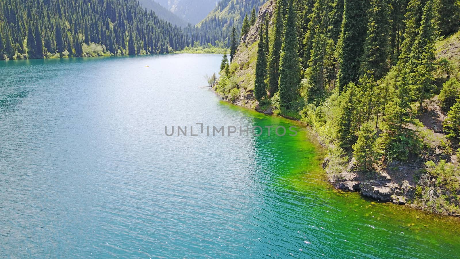 Kolsay lake among green hills and mountains. The mountain lake is surrounded by green forest, tall coniferous trees, grass and bushes. Clean water is like a mirror. Tourists swim on boats. Kazakhstan