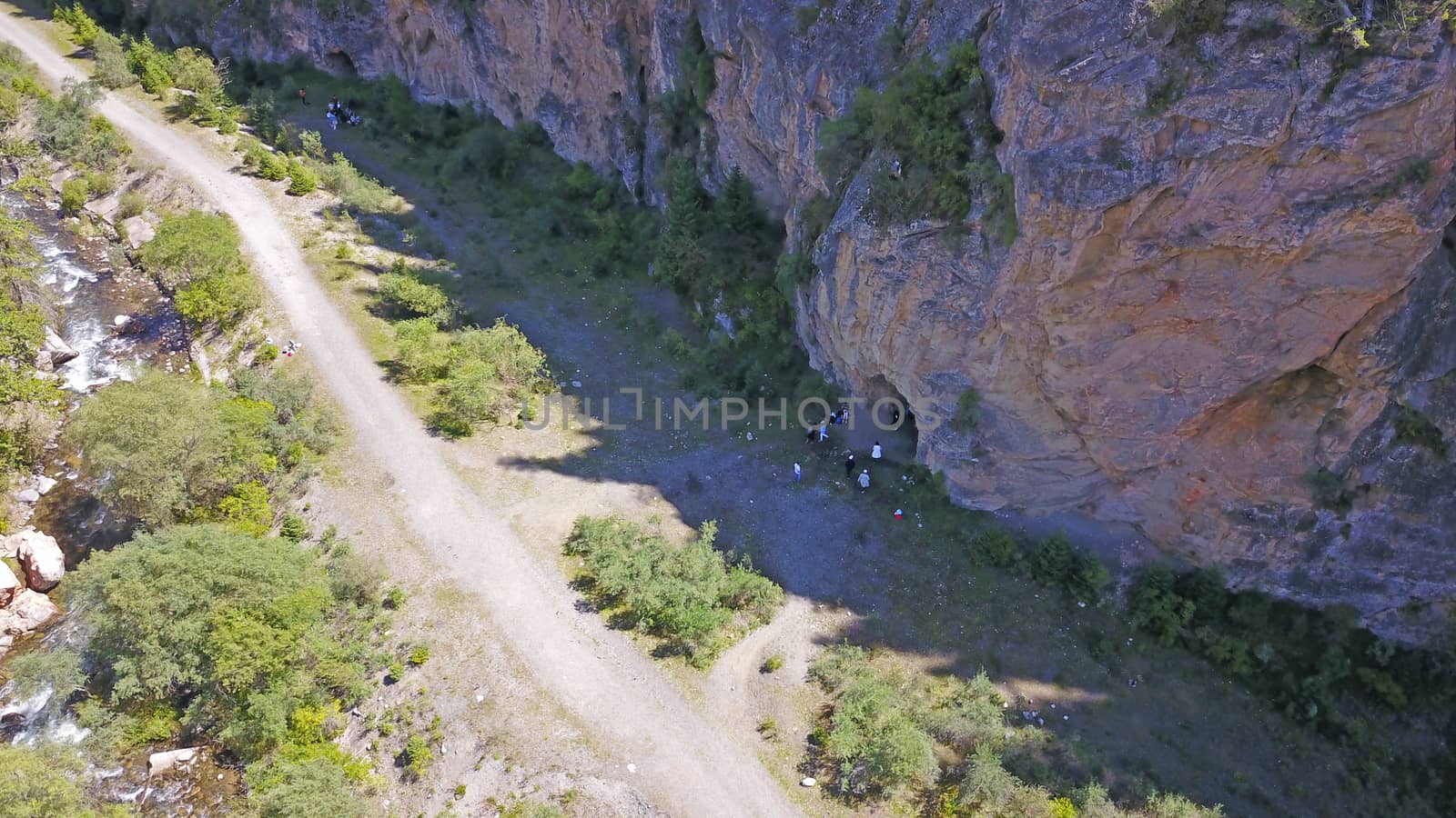 Top view of the cave, forest, rocks and road. A group of tourists walking along the trail. There is a huge cave in the rock. Green coniferous forest, trees and grass grow on the rocks. The river runs.