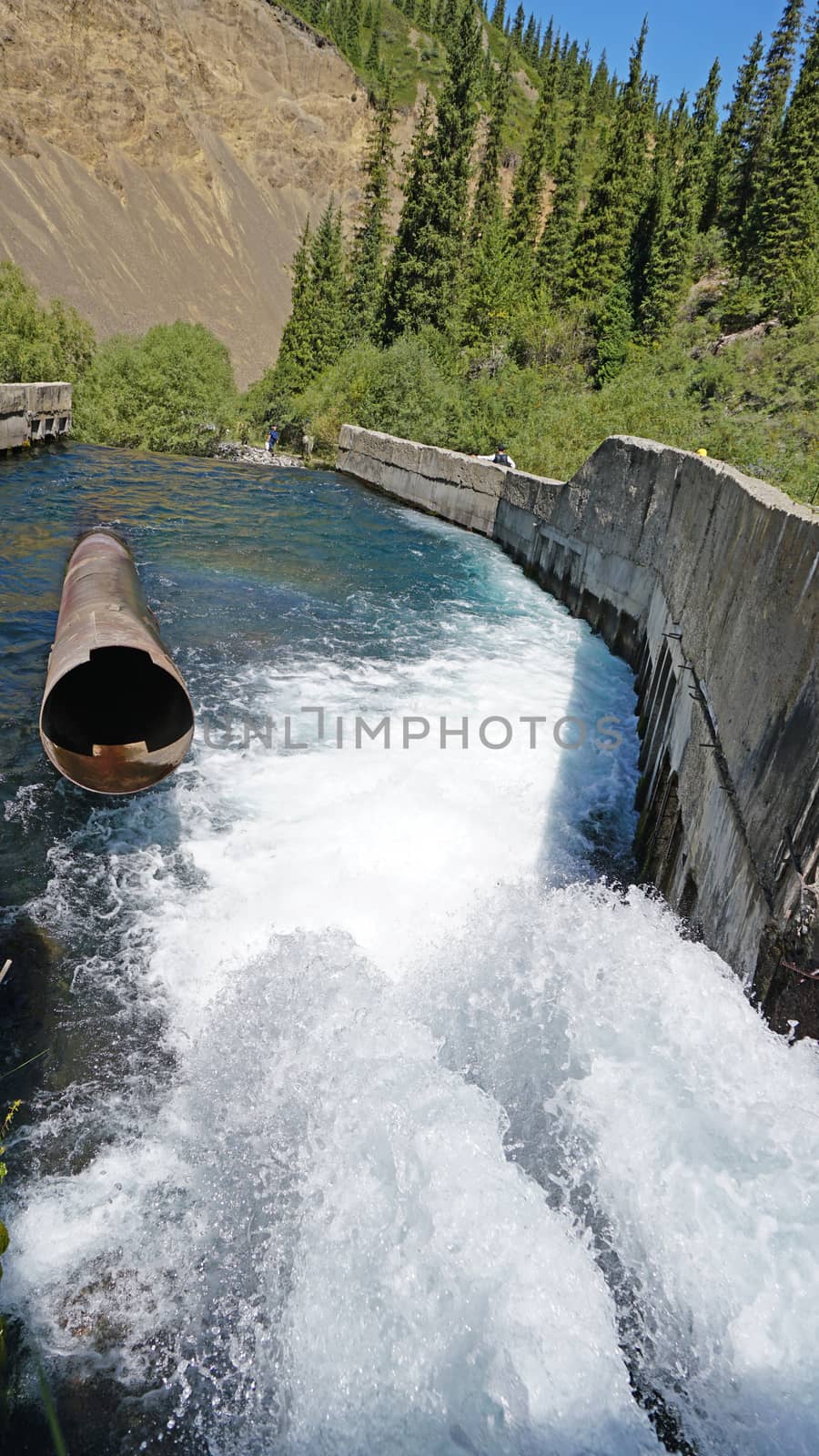 Draining the water pressure into the river. The water flows like a waterfall and flows into the river. There are concrete walls around the edges, and iron pipes are visible. Trees grow in the distance