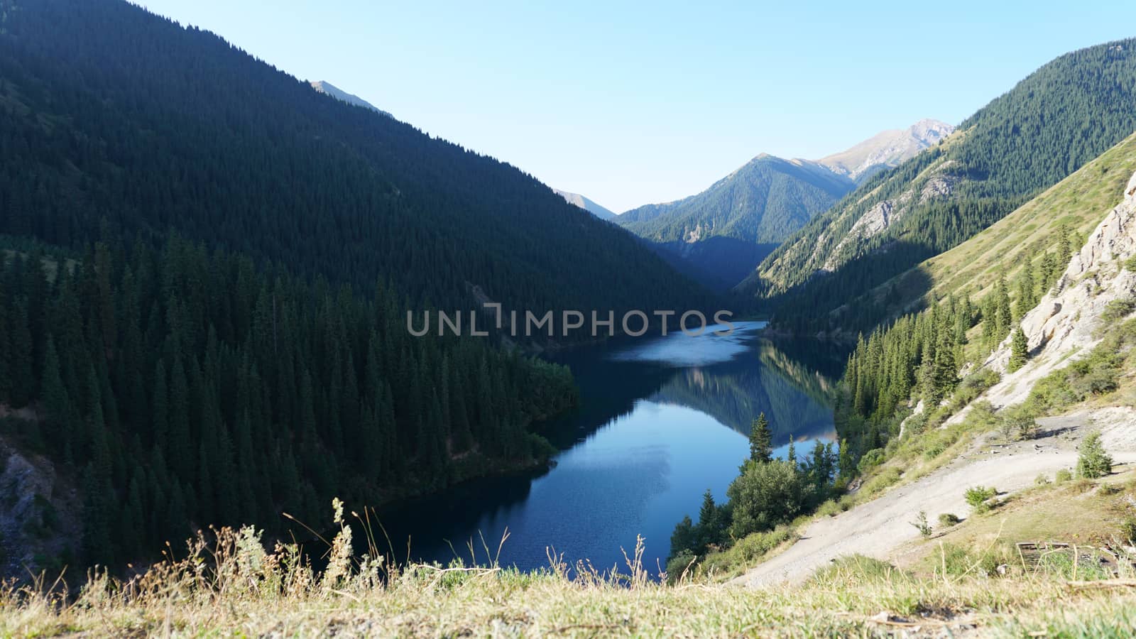 Kolsay lake among green hills and mountains. The mountain lake is surrounded by green forest, tall coniferous trees, grass and bushes. Clean water is like a mirror. Tourists swim on boats. Kazakhstan