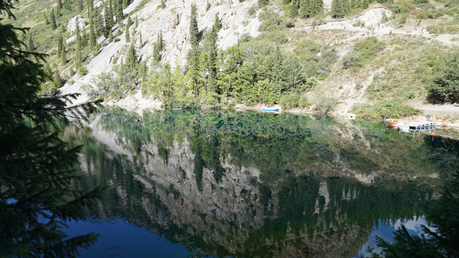 Kolsay lake among green hills and mountains. The mountain lake is surrounded by green forest, tall coniferous trees, grass and bushes. Clean water is like a mirror. Tourists swim on boats. Kazakhstan