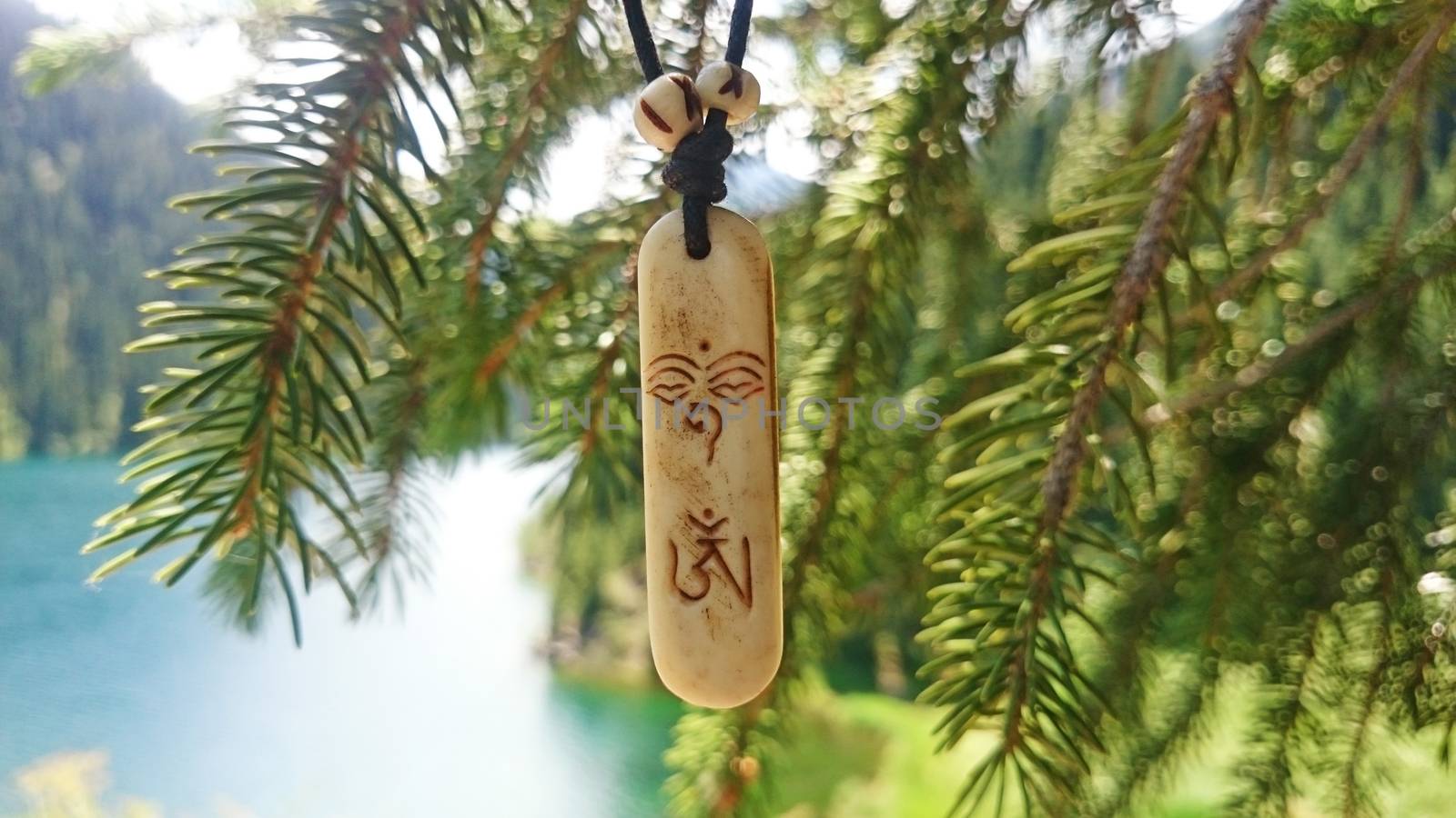 Macro symbol OM (AUM). A locket with OM symbol. The background is blurred. Coniferous trees, a lake, fir branches, blue and green water of the lake are visible. Mountain lake. Kazakhstan, Almaty.