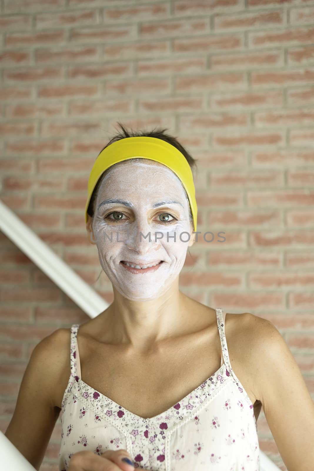 Relaxed woman applies mineral clay mask on face for rejuvenation, has headband on head