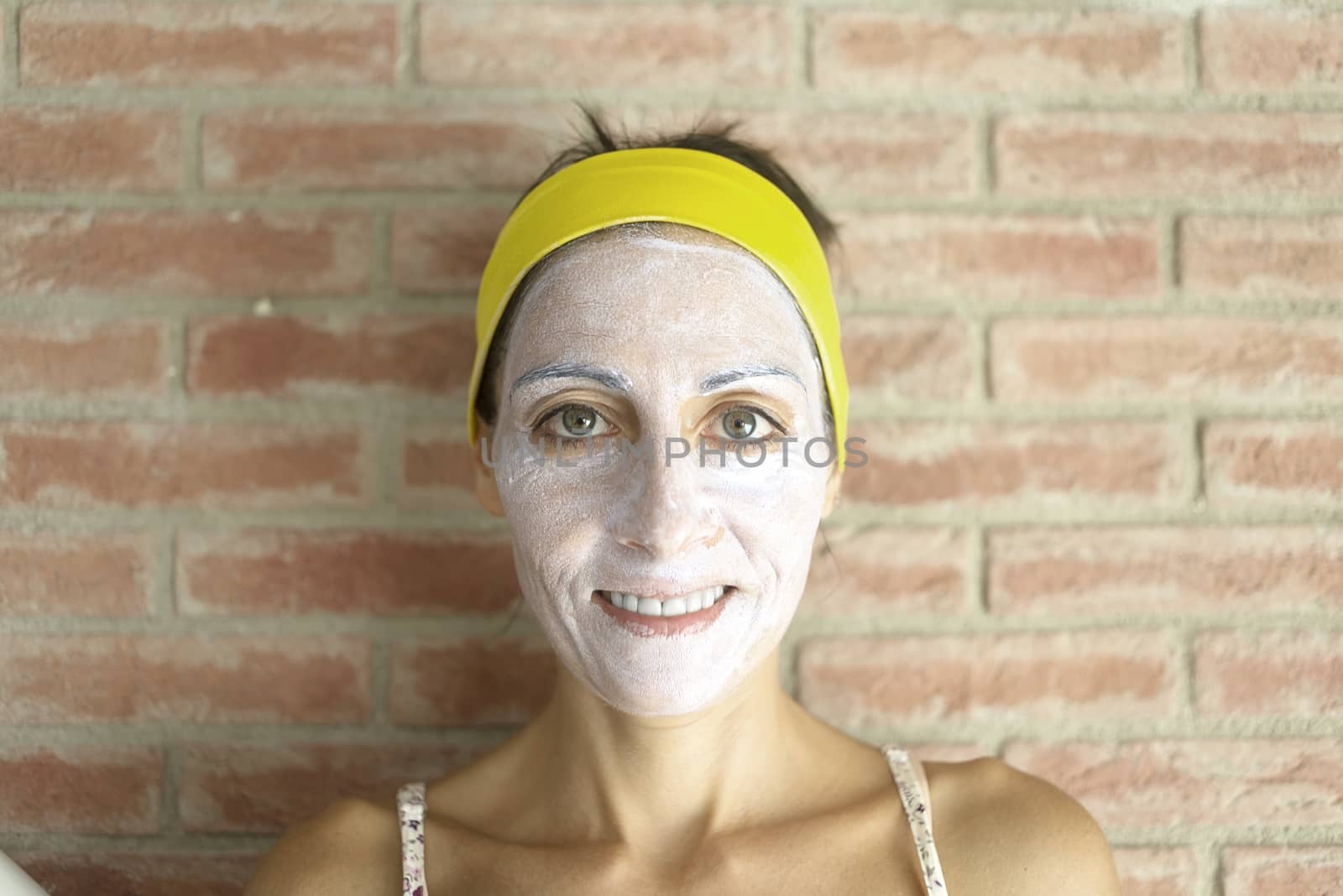 Relaxed woman applies mineral clay mask on face for rejuvenation, has headband on head