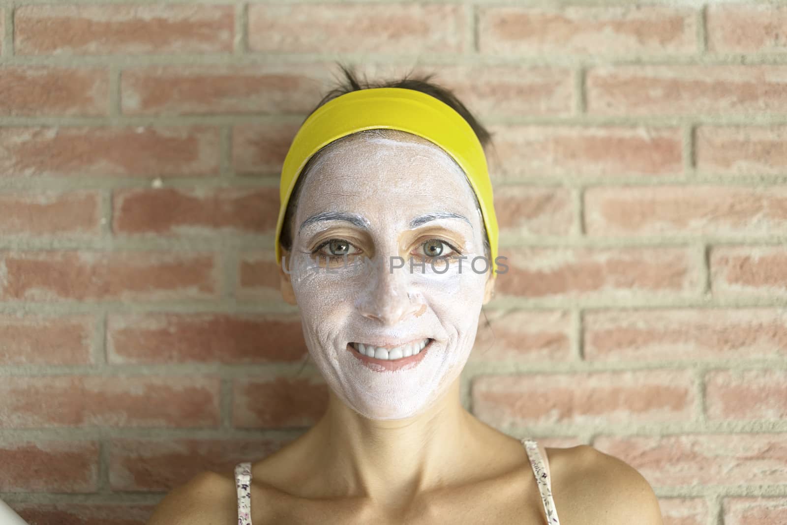 Relaxed woman applies mineral clay mask on face for rejuvenation, has headband on head
