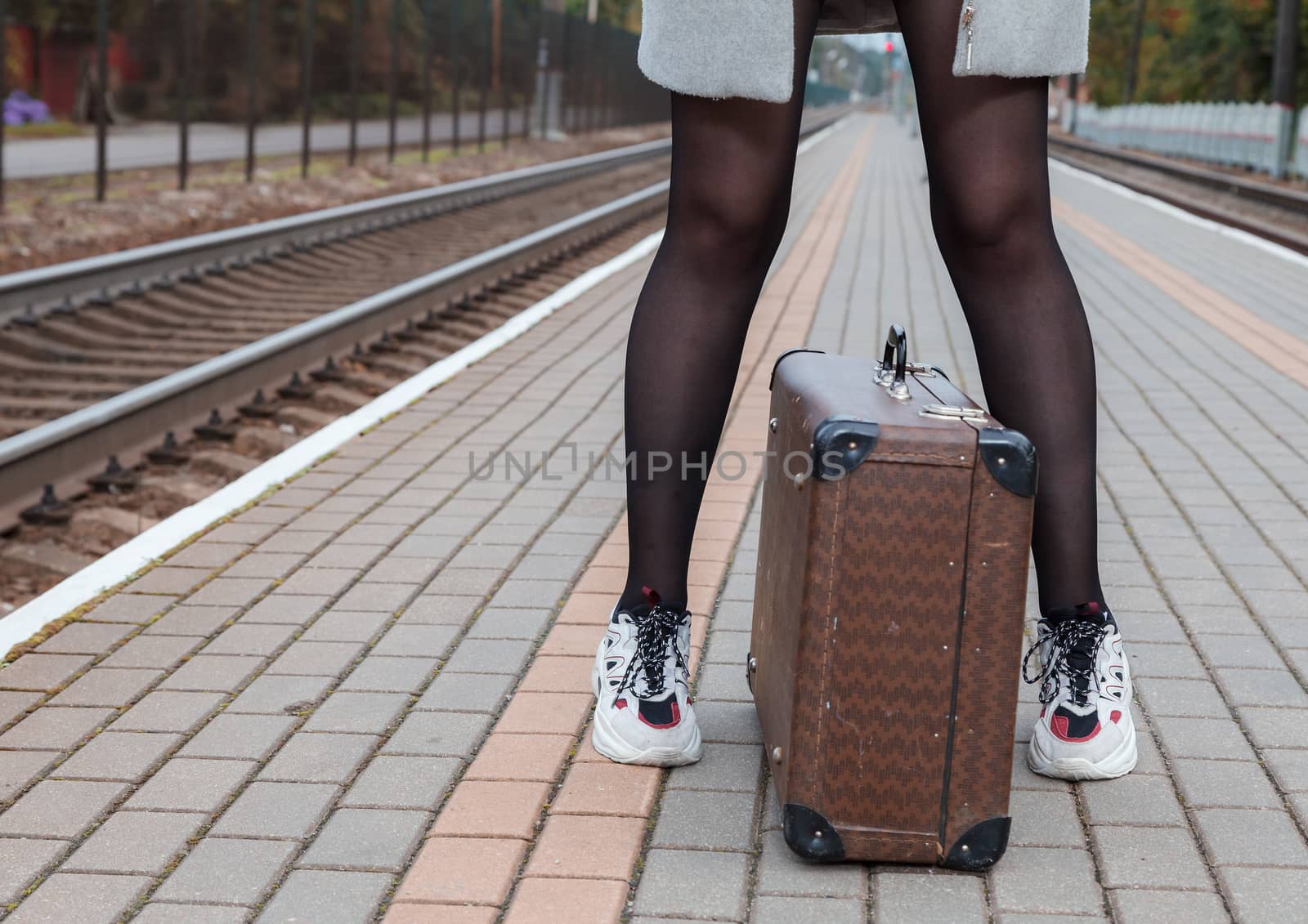 woman in a gray coat and with a suitcase standing at the railway station on an autumn day. legs closeup