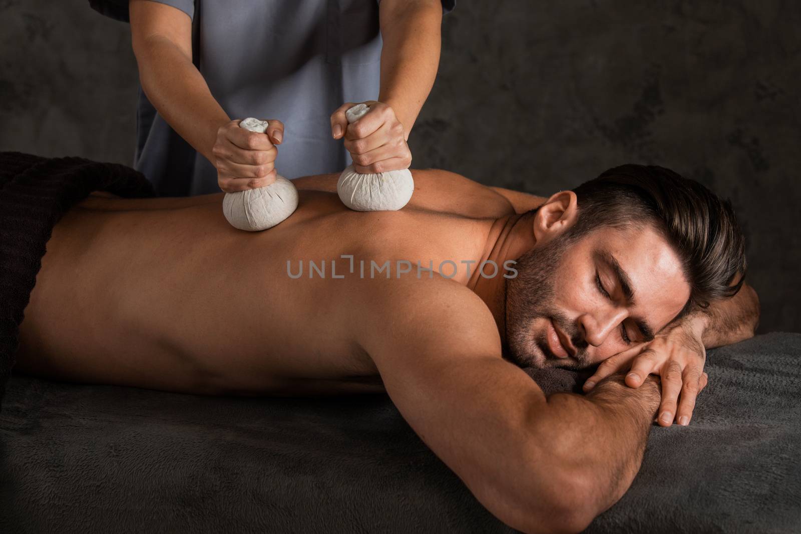 A good-looking man getting a back massage lying down at spa, pouches filled with dry herbs are brushed over body close up