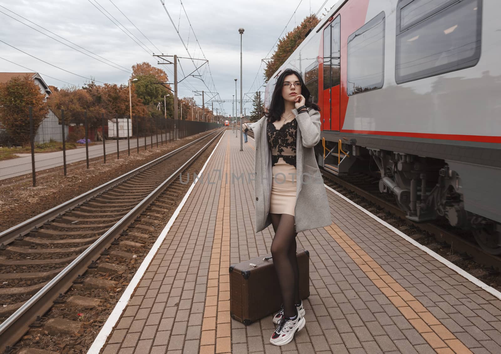 lonely young girl in gray coat with suitcase standing at railway station on gloomy autumn day