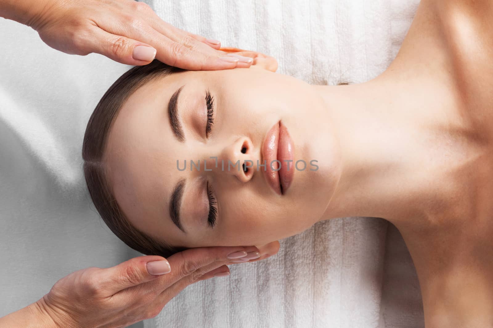 Facial massage, top view of beautiful young woman lying on back and keeping her eyes closed while massage therapist massaging her face