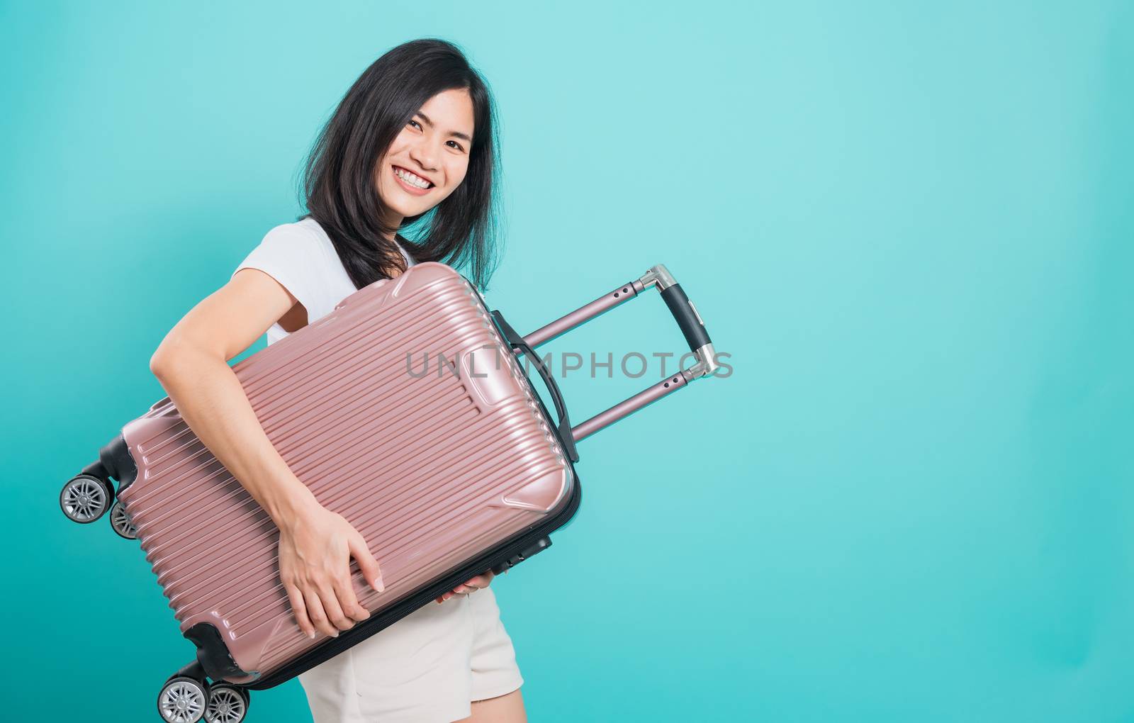 Portrait happy Asian beautiful young woman standing wear white t-shirt, holidays travel concept, her hugging suitcase bag and looking to camera, shoot photo in a studio on a blue background