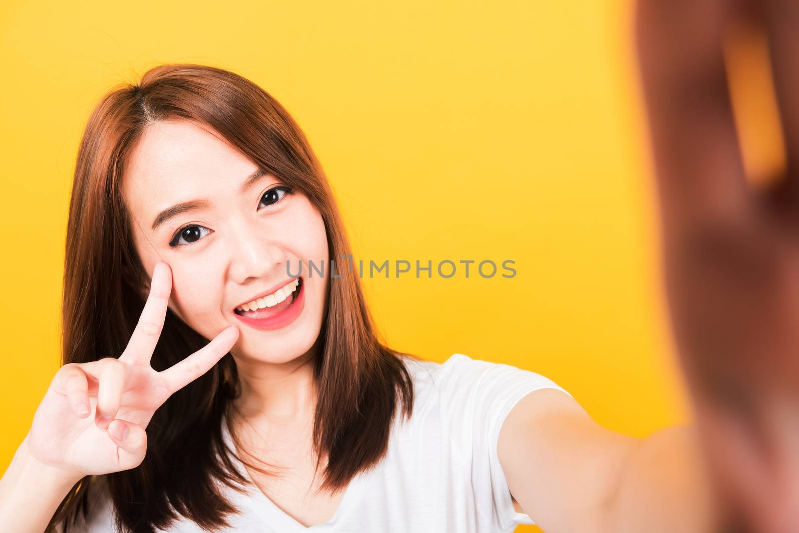 Asian happy portrait beautiful cute young woman teen smiling standing wear t-shirt making selfie photo, video call on smartphone looking camera isolated, studio shot yellow background with copy space