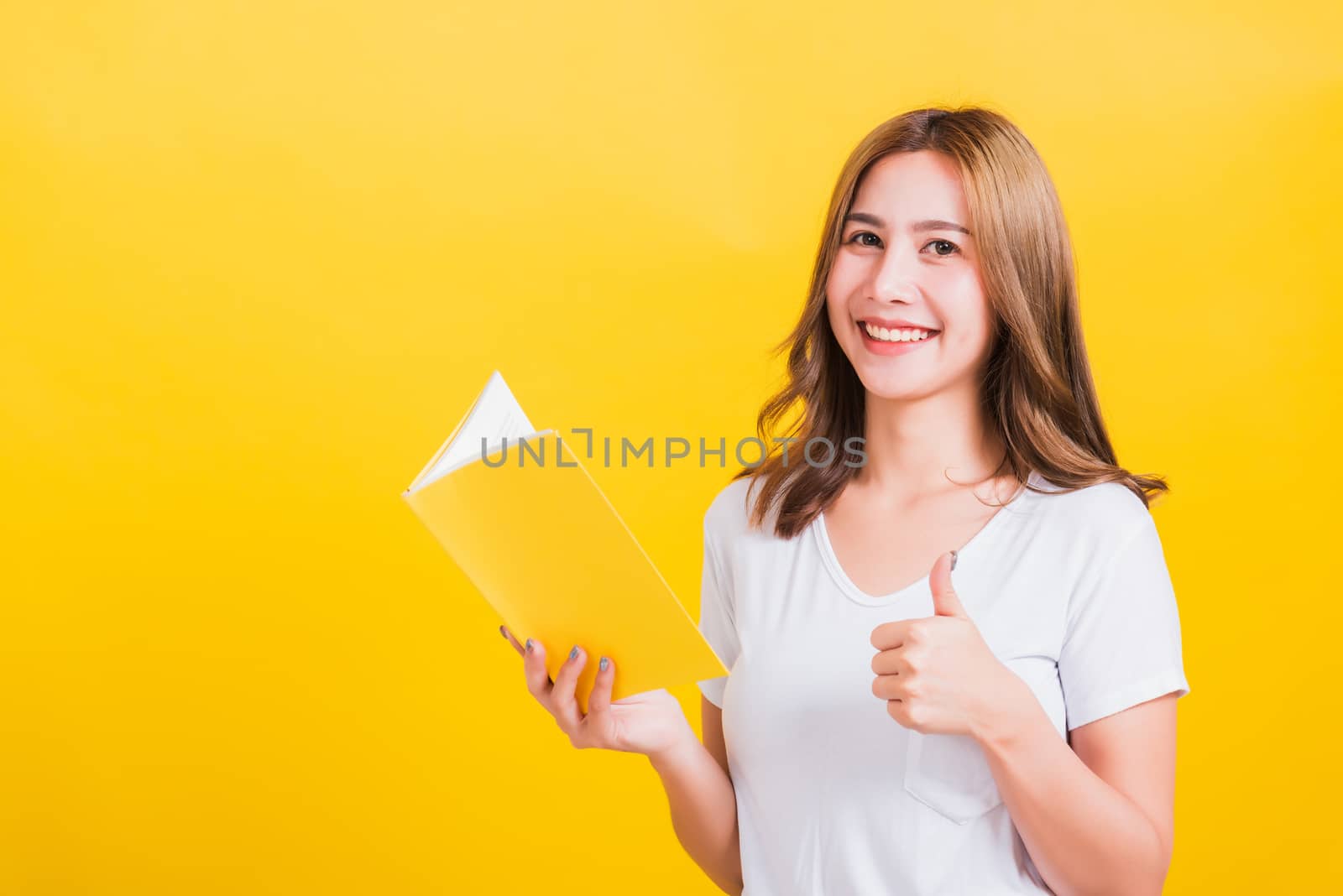 Portrait Asian Thai beautiful happy young lifestyle woman stands holding yellow book or diary she show thumb up finger and looking to camera, studio shot isolated on yellow background, with copy space