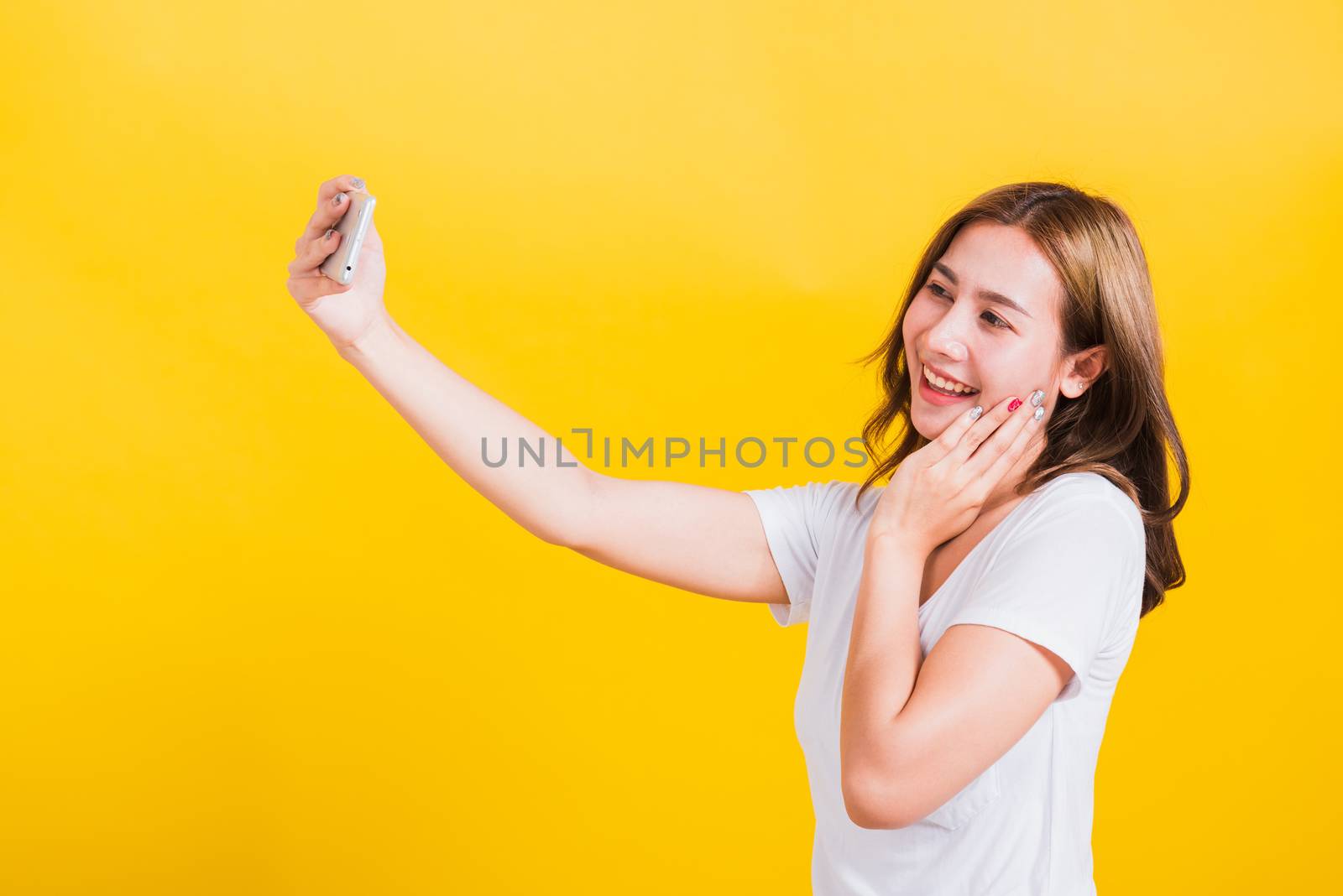 Asian Thai portrait happy beautiful cute young woman smiling wear t-shirt making selfie photo or video call on smartphone looking the phone, studio shot isolated yellow background with copy space