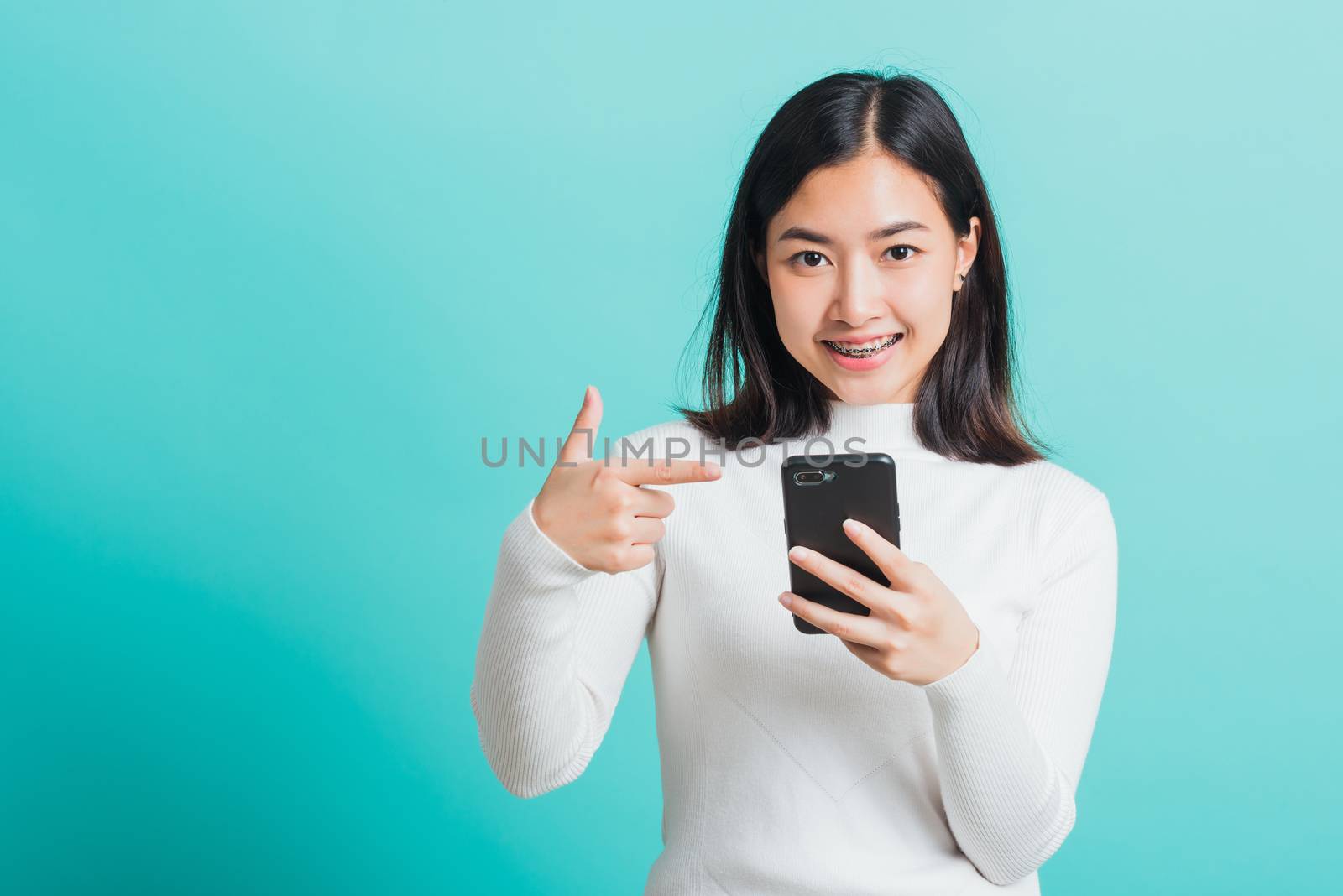 Beautiful Asian woman smile she pointing finger to a smartphone, female excited cheerful after received promotion her point finger on mobile phone isolated on a blue background, Technology concept
