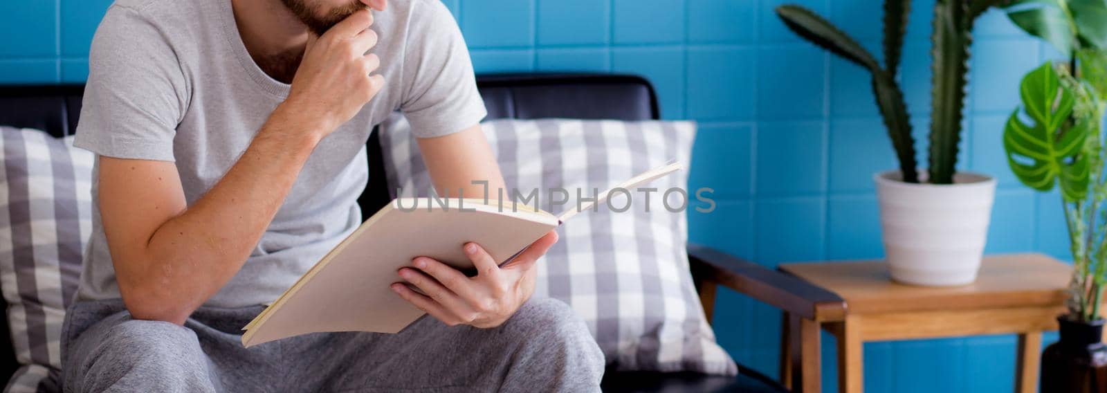 Young man reading a book sitting on sofa for rest is hobby in living room, stay home for leisure, lifestyle of new normal for social distracting, male learn and study in interesting in spare time. by nnudoo