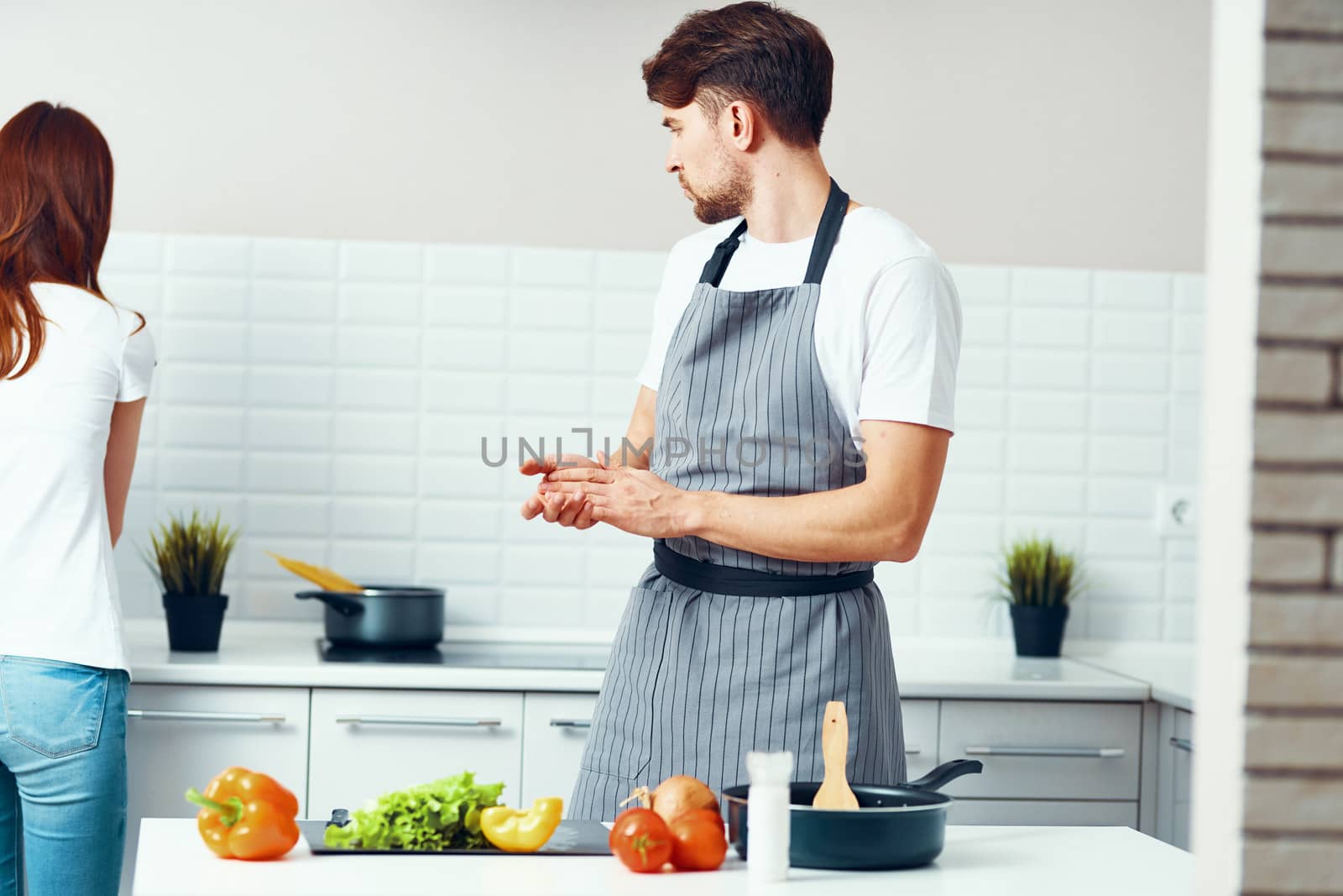 chef in an apron in the kitchen with fresh vegetables and a woman in jeans in the background