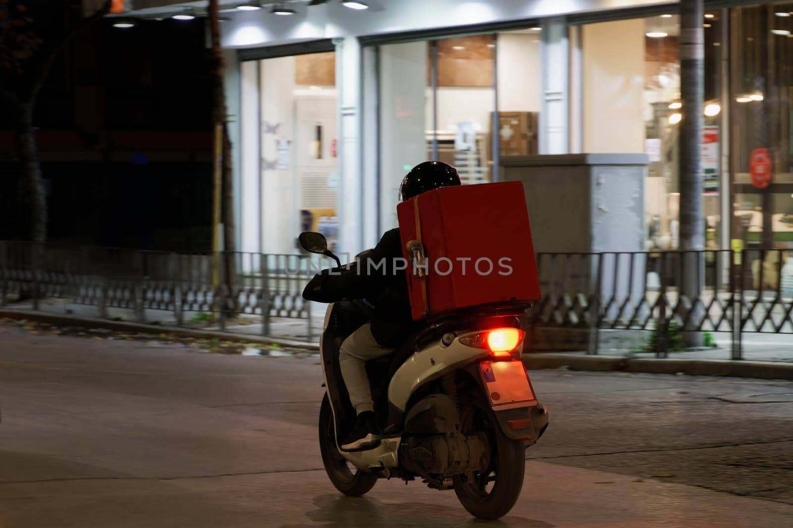 Illuminated view of male with helmet riding motorcycle to deliver take away food contained on a red box in Thessaloniki, Greece.