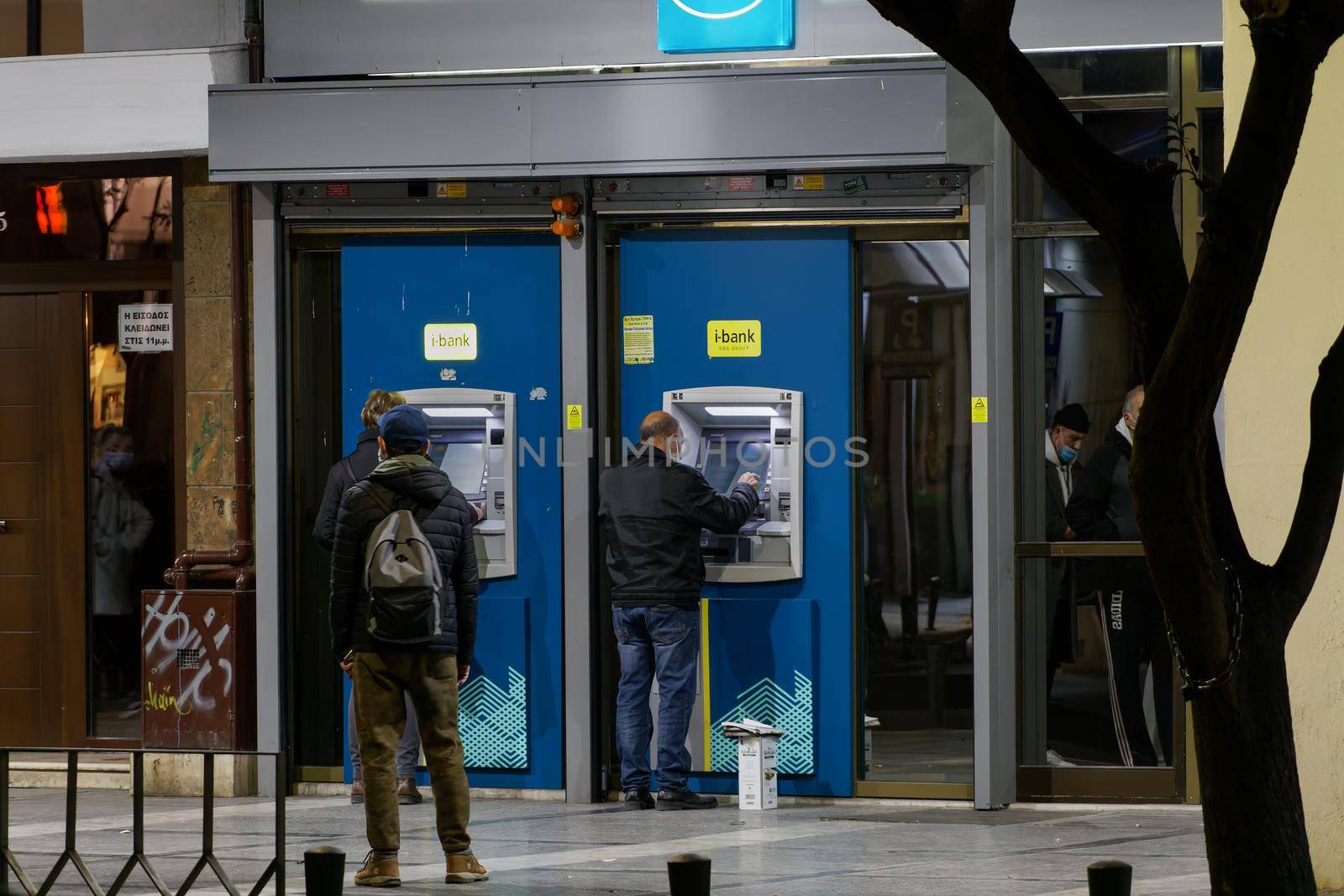 Illuminated night view of unidentified people using Hellenic 24-hour cash machines outside a branch of National Bank.