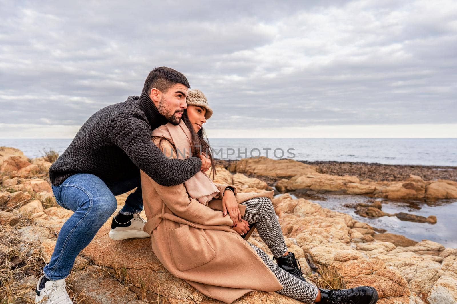 Beautiful heterosexual mixed race couple outdoor live a tenderness moment sitting on sea resort rocks - Caucasian handsome guy embracing his Hispanic girlfriend in the nature with orange color mood by robbyfontanesi
