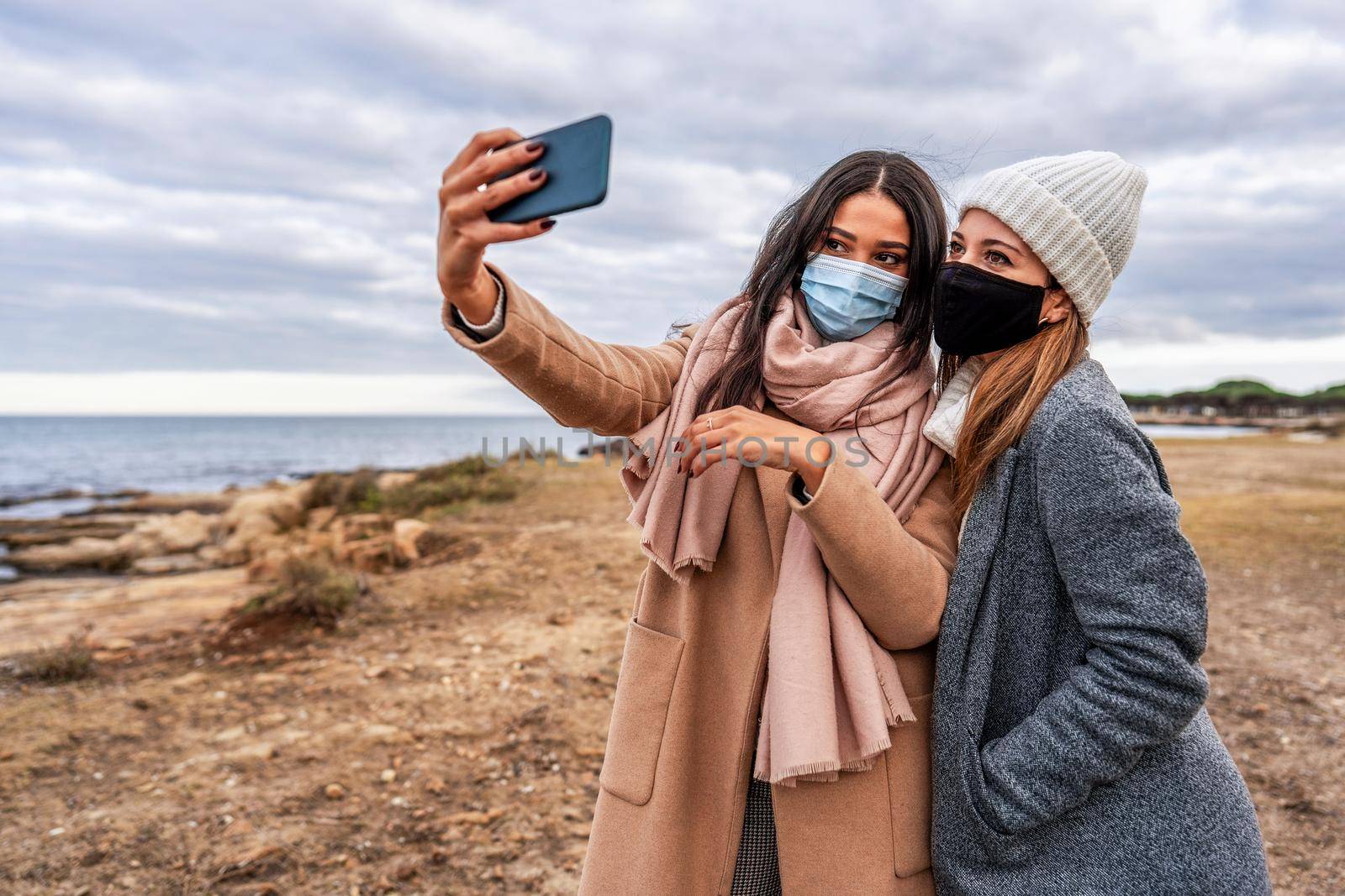 Young mixed race female LGBTQ couple making selfie with smartphone in sea resort in safe protection mask - Hispanic beautiful woman using tech to sharing photo with her lesbian girlfriend on social