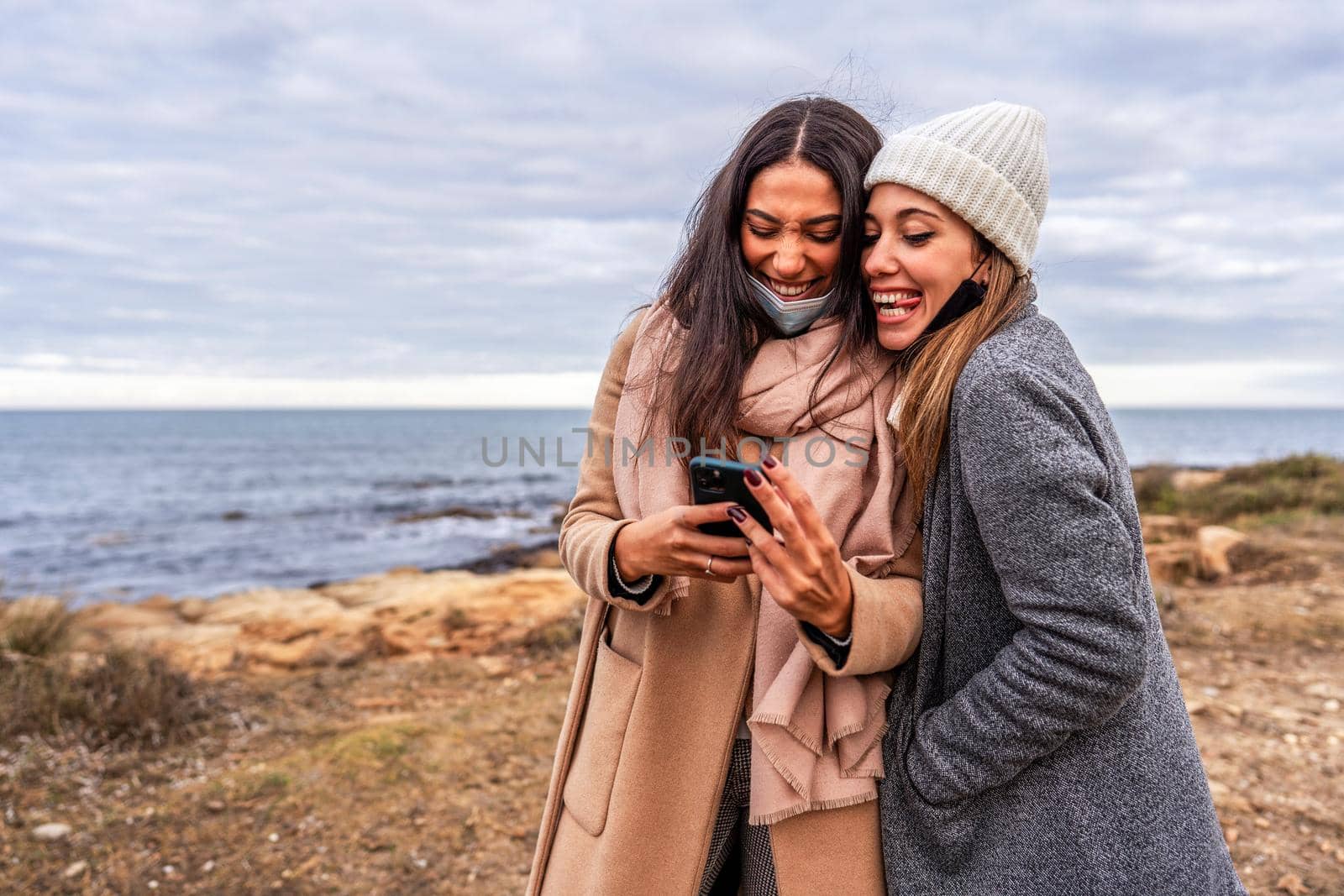 Young mixed race female LGBTQ couple having fun making faces with tongue out laughing watching photos on smartphone - Two beautiful lesbian women joking with internet outdoor in sea winter vacation by robbyfontanesi