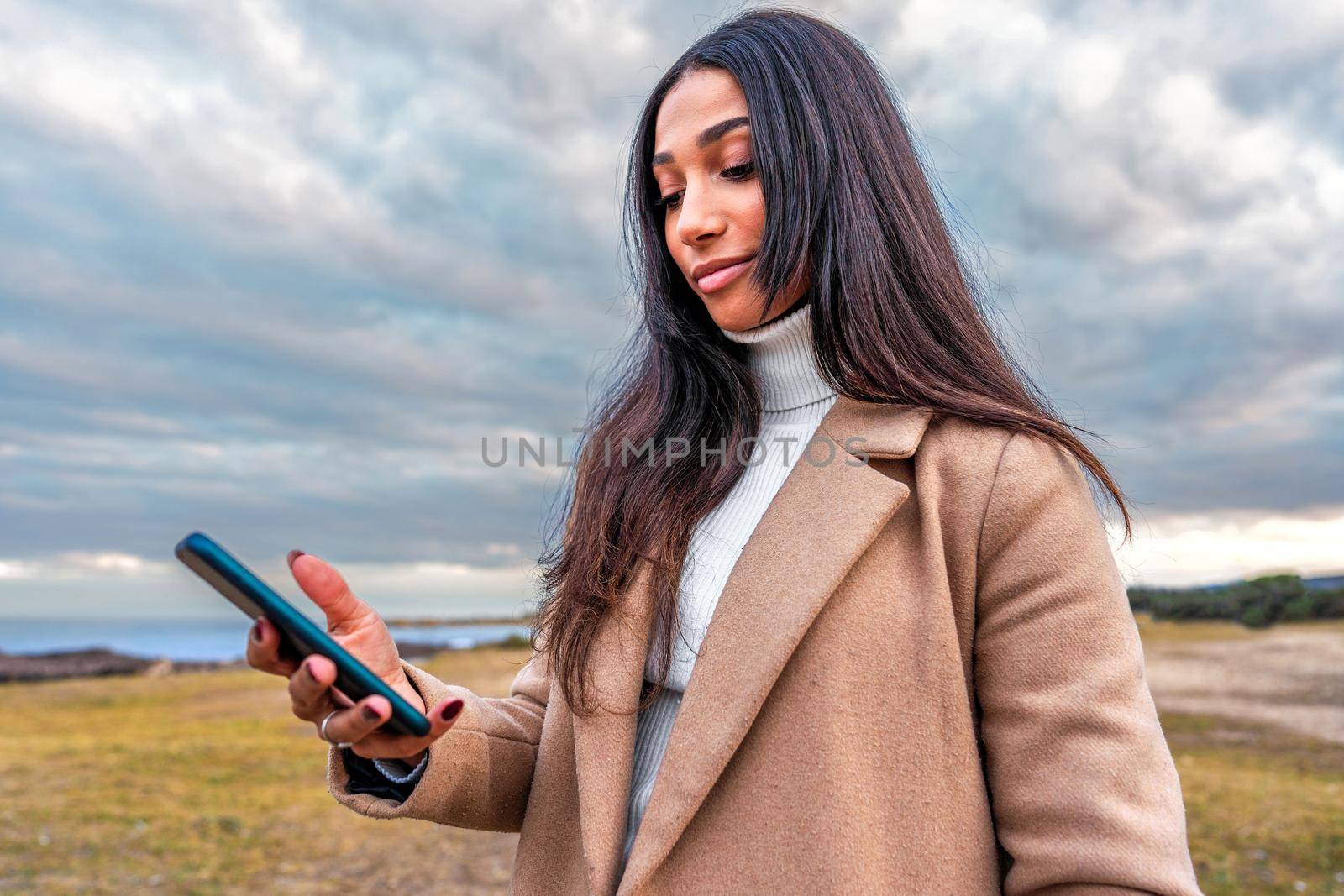 Bottom view portrait of beautiful young Hispanic woman with black long hair standing outdoor in sea resort watching smartphone in hand - Fall-winter colors filter photography with dramatic cloudy sky by robbyfontanesi